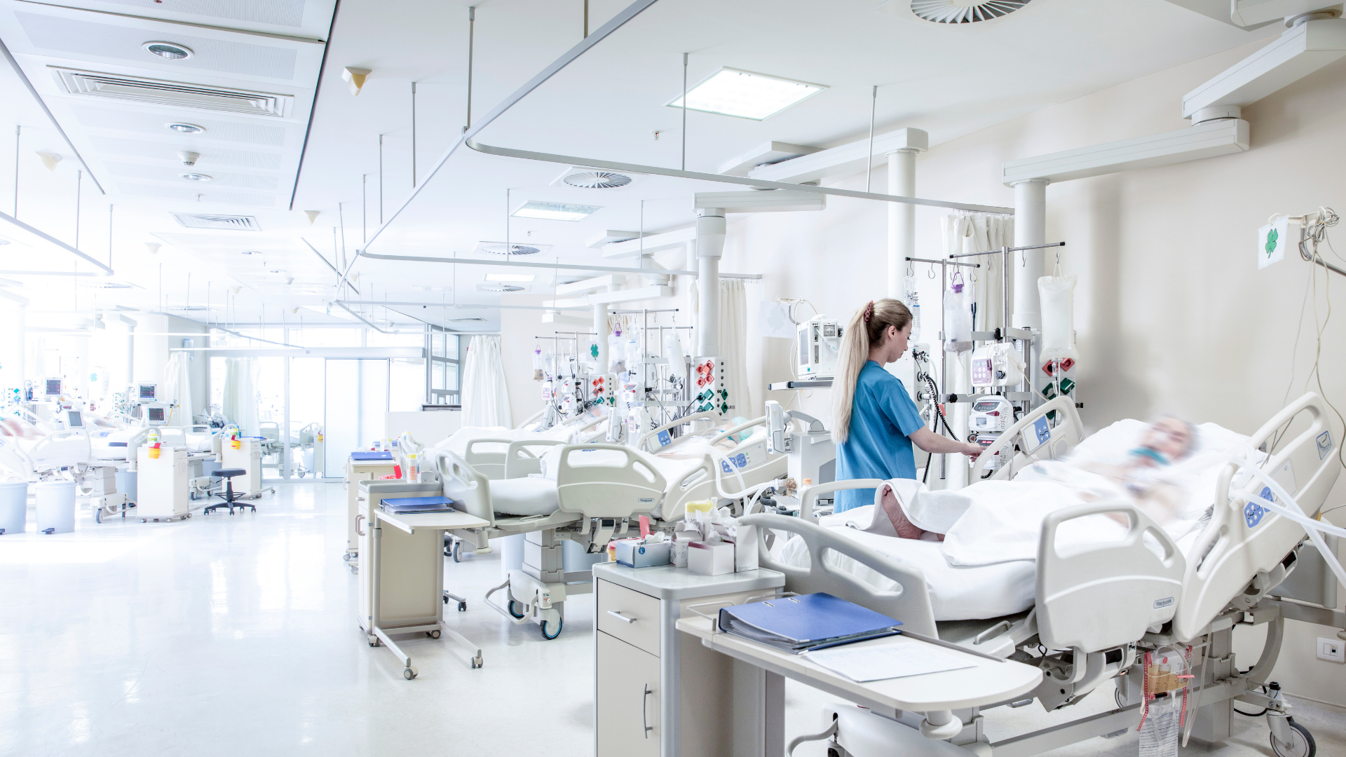 A hospital ward with multiple empty beds, medical equipment, and a nurse attending to a patient lying in a bed.