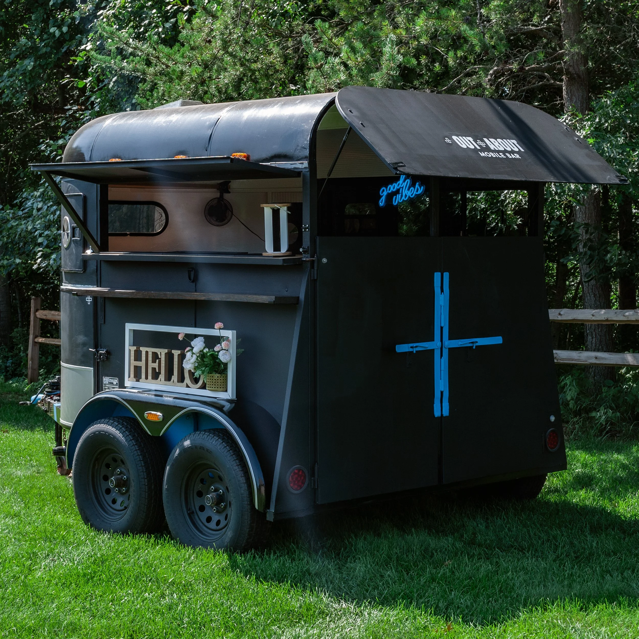 Black food trailer parked on green grass with trees in the background. The trailer has a sign that reads 'Out About Mobile Bar' and a small neon sign that says 'Good Vibes'.