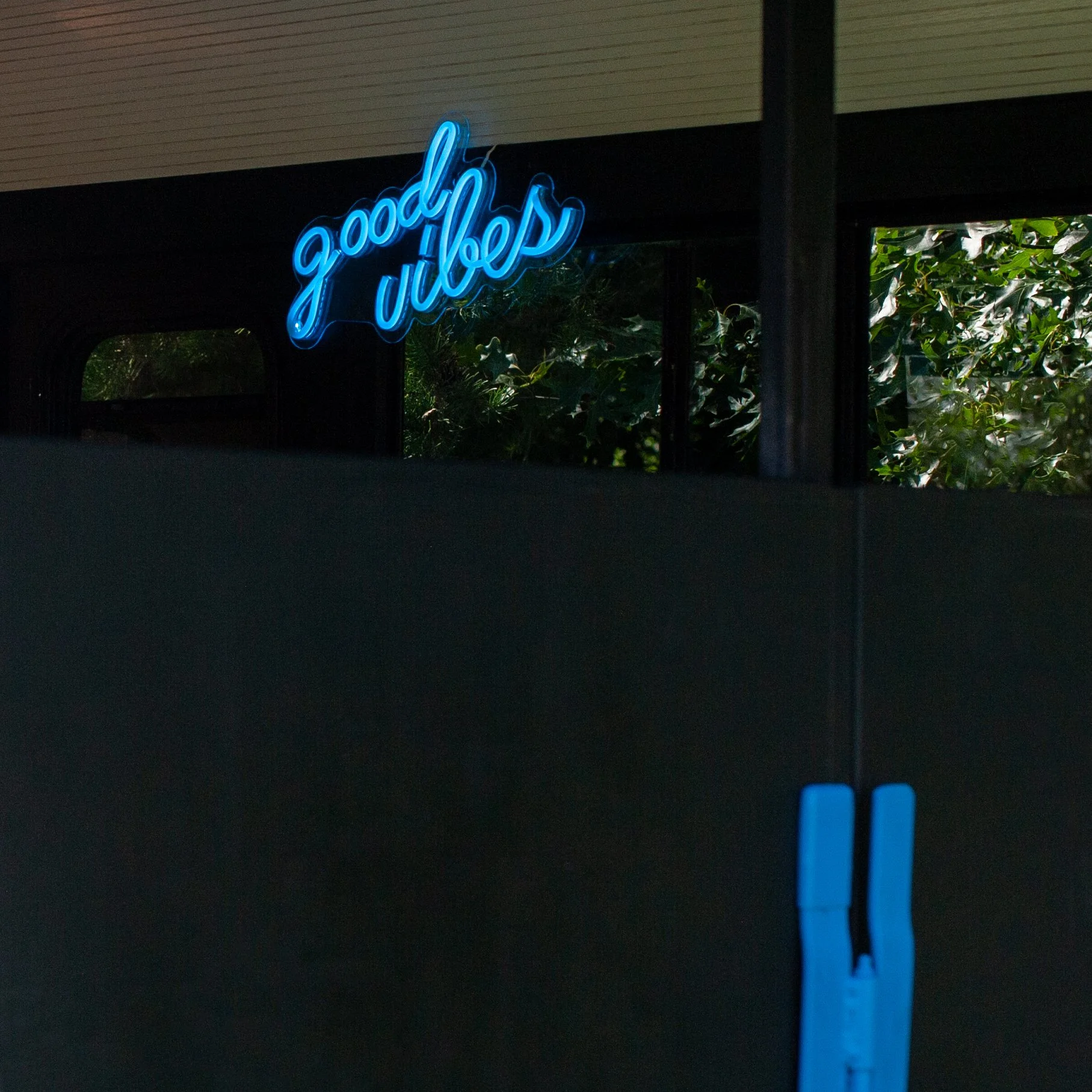 A neon sign that reads 'good vibes' in blue, with a background of dark walls, a window showing greenery outside, and a structure at the bottom right.
