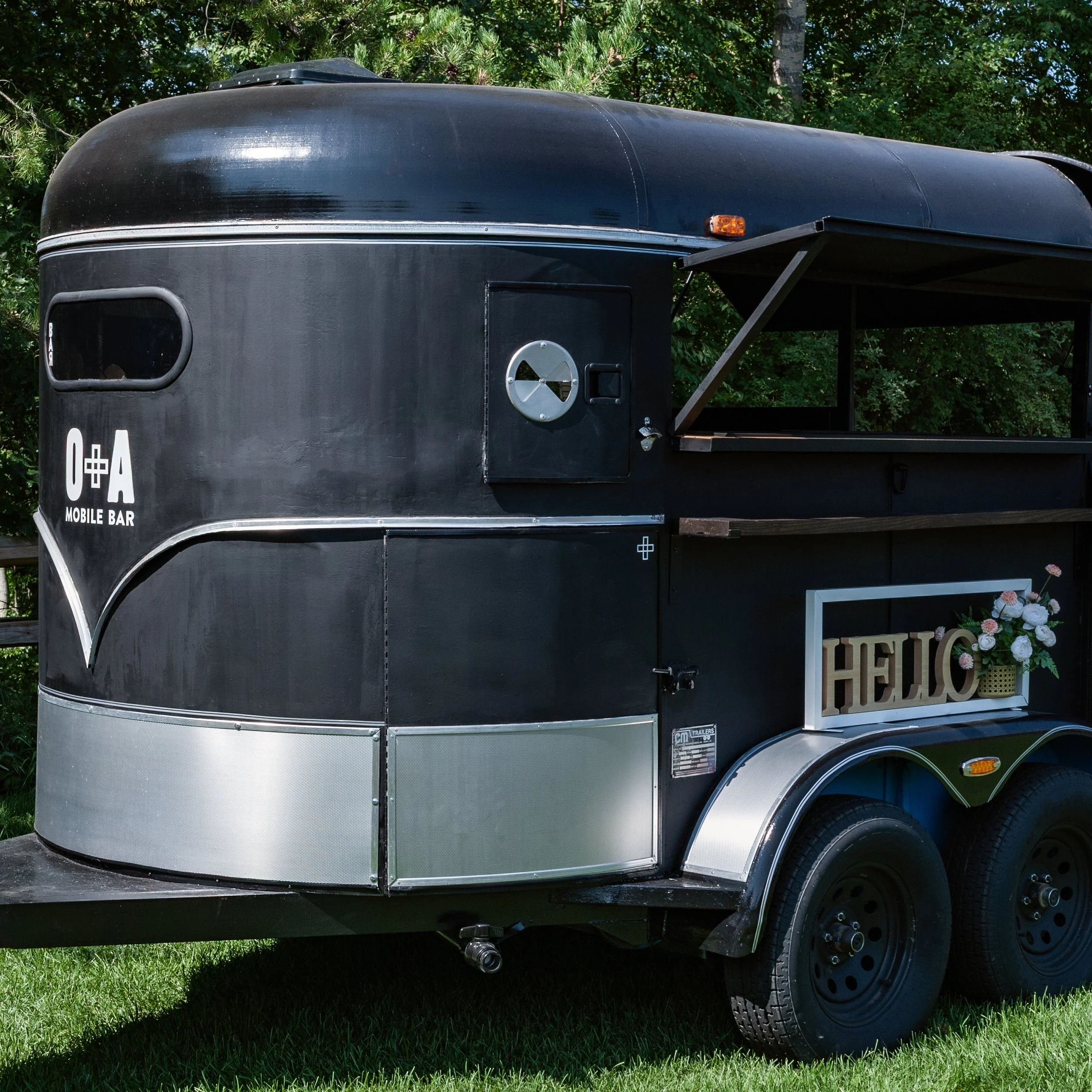 Black food trailer with a window, silver trim, and a small vent on top, parked on grass with trees in the background.