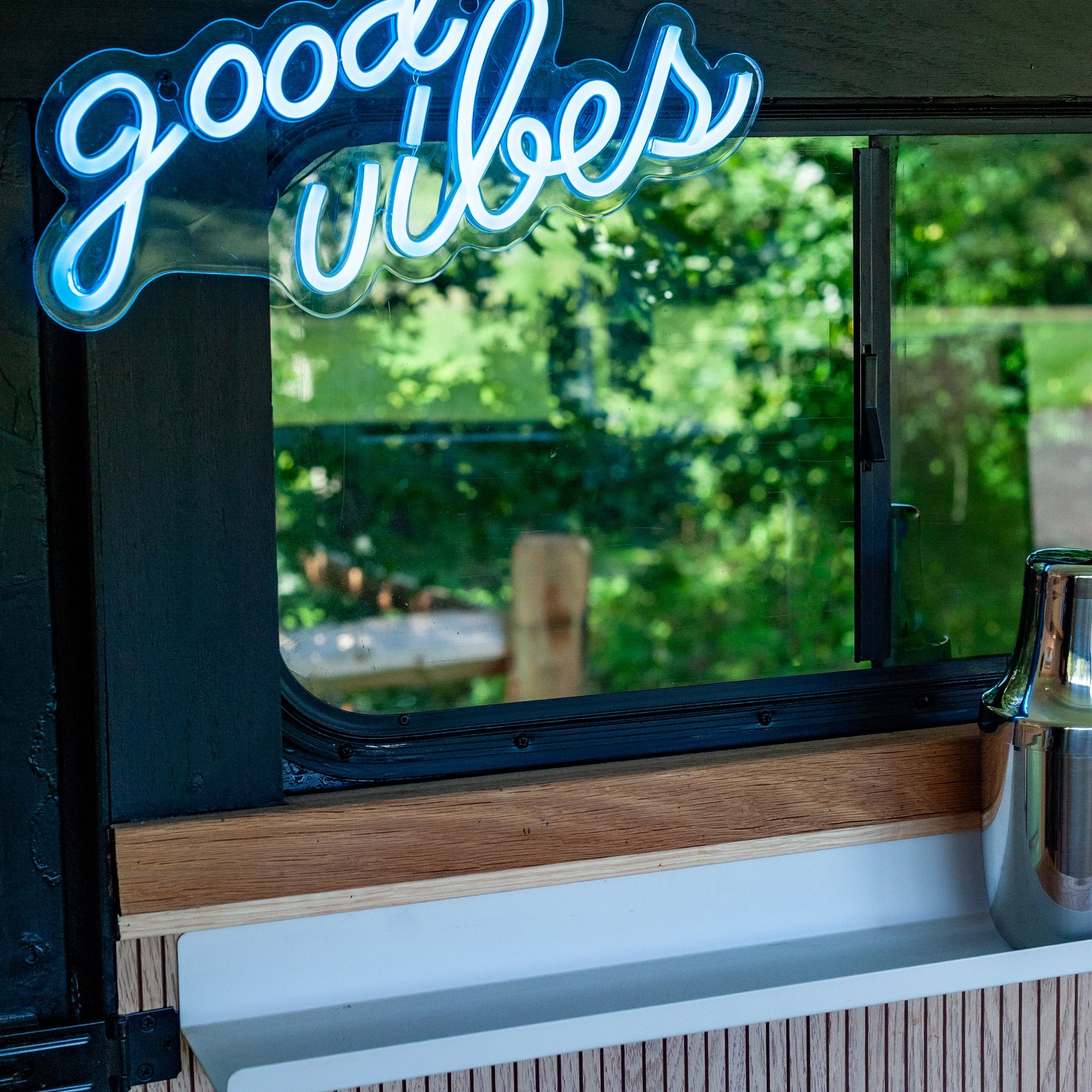 Neon sign that says "Good Vibes" in blue, mounted on a window with a reflection of trees outside.