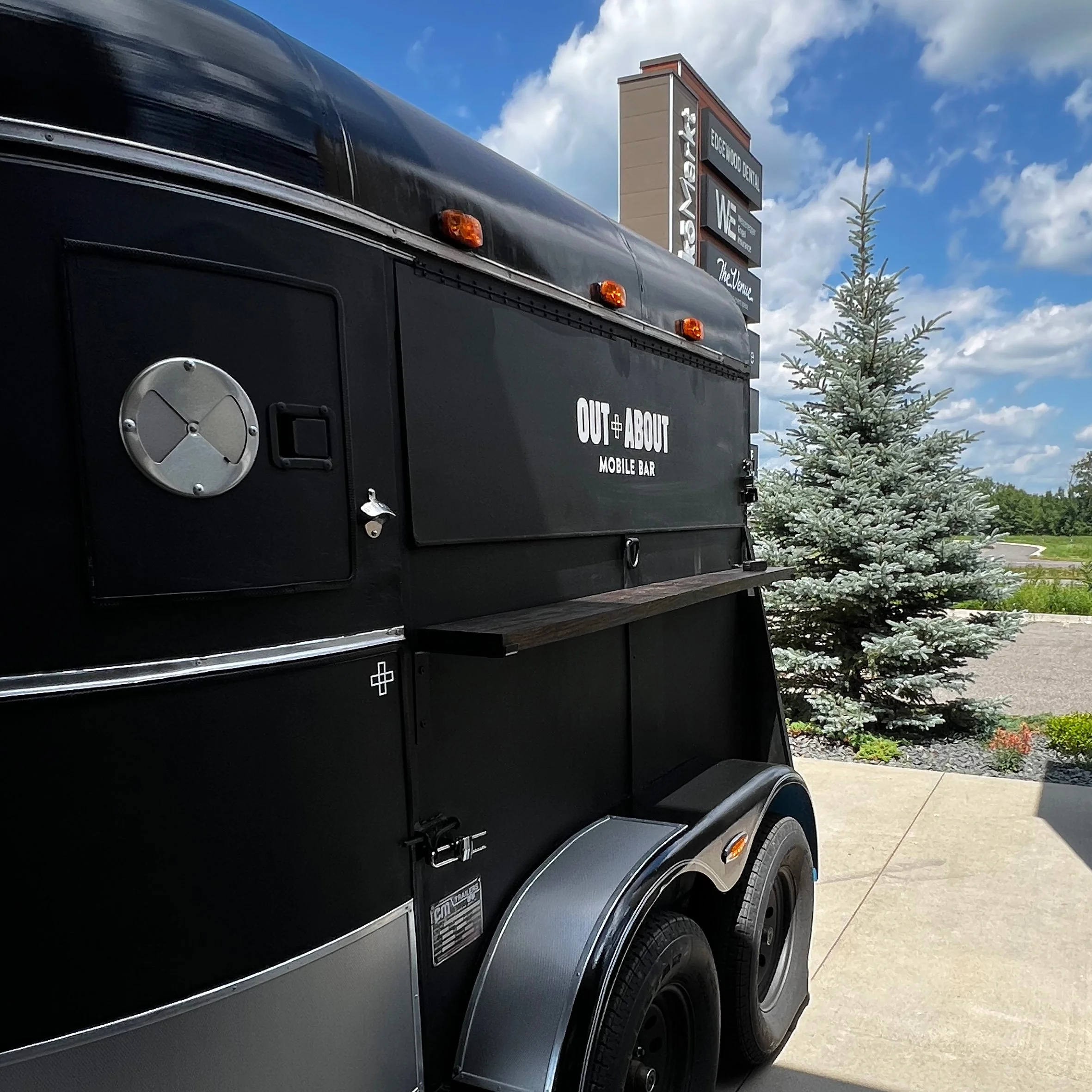 Close-up of a black food truck with the words 'Out and About' and 'Mobile Bar' written on it, parked next to a tree with a blue sky in the background.