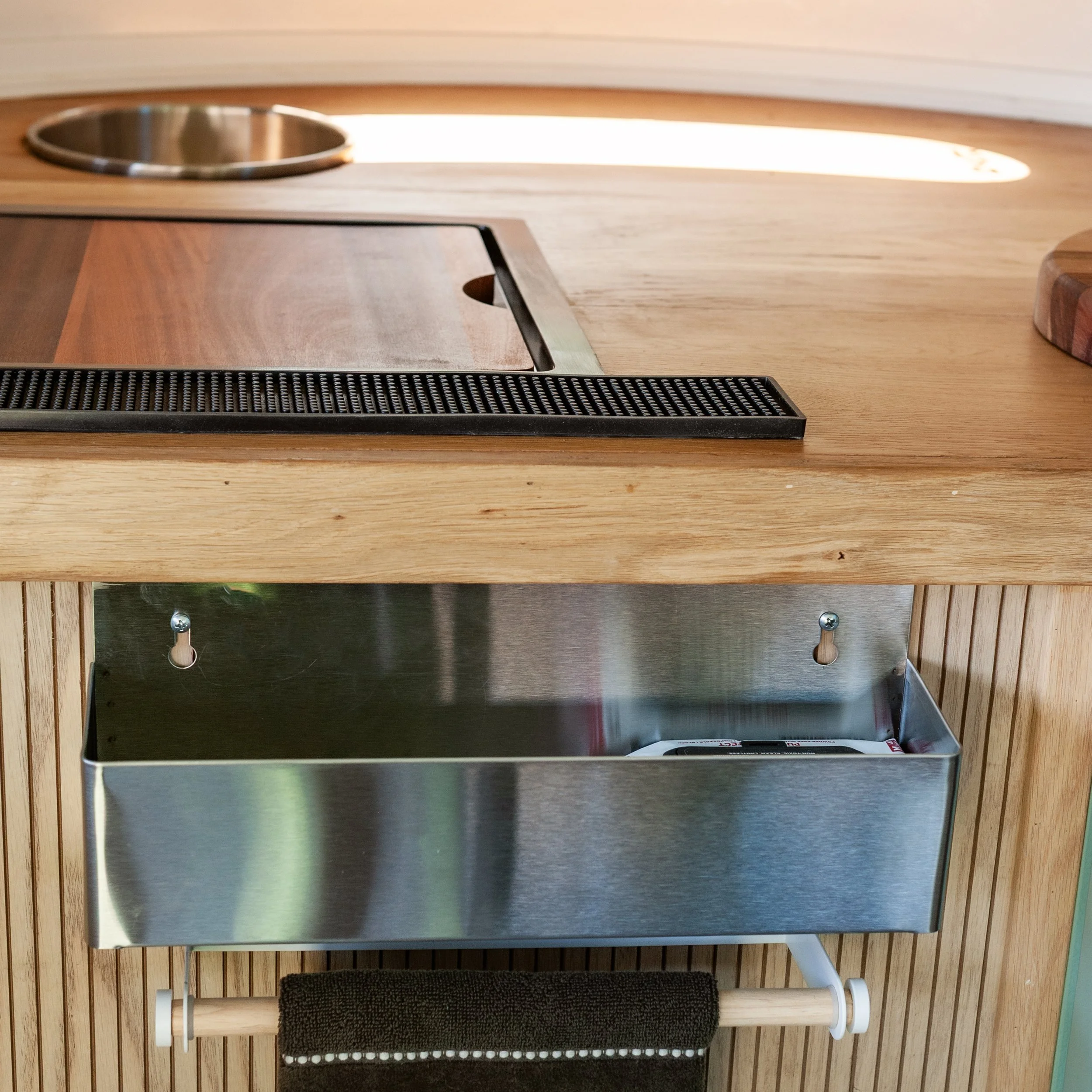 A modern wooden countertop oven with a black metal front panel and a dark towel hanging on a wooden towel rack below. The oven is positioned against a wall with vertical wood paneling.