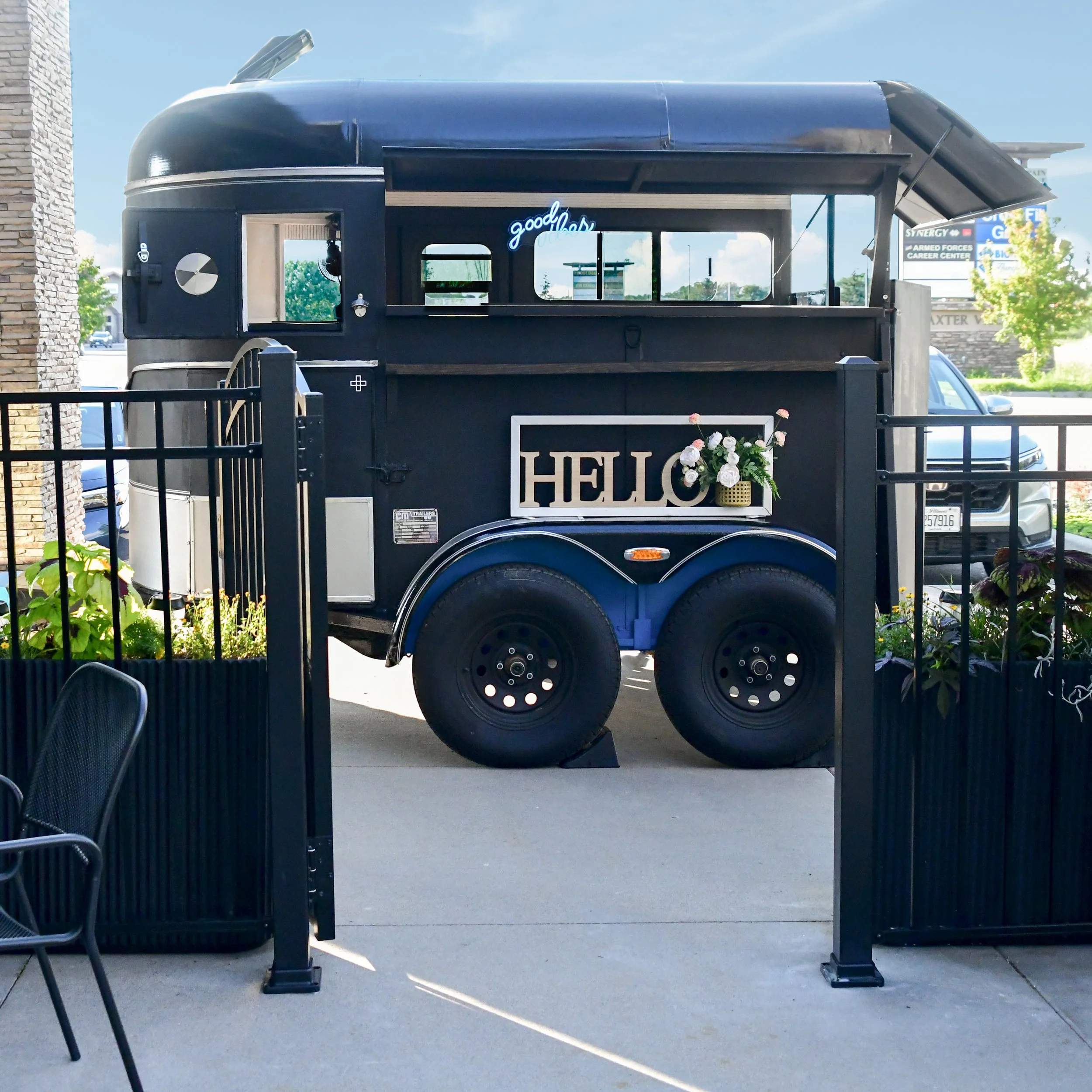 A food truck with a dark exterior featuring a neon sign that says 'good vibes' and a sign that says 'HELLO' decorated with white and pink flowers, parked on a sunny day near a sidewalk with a black fence.