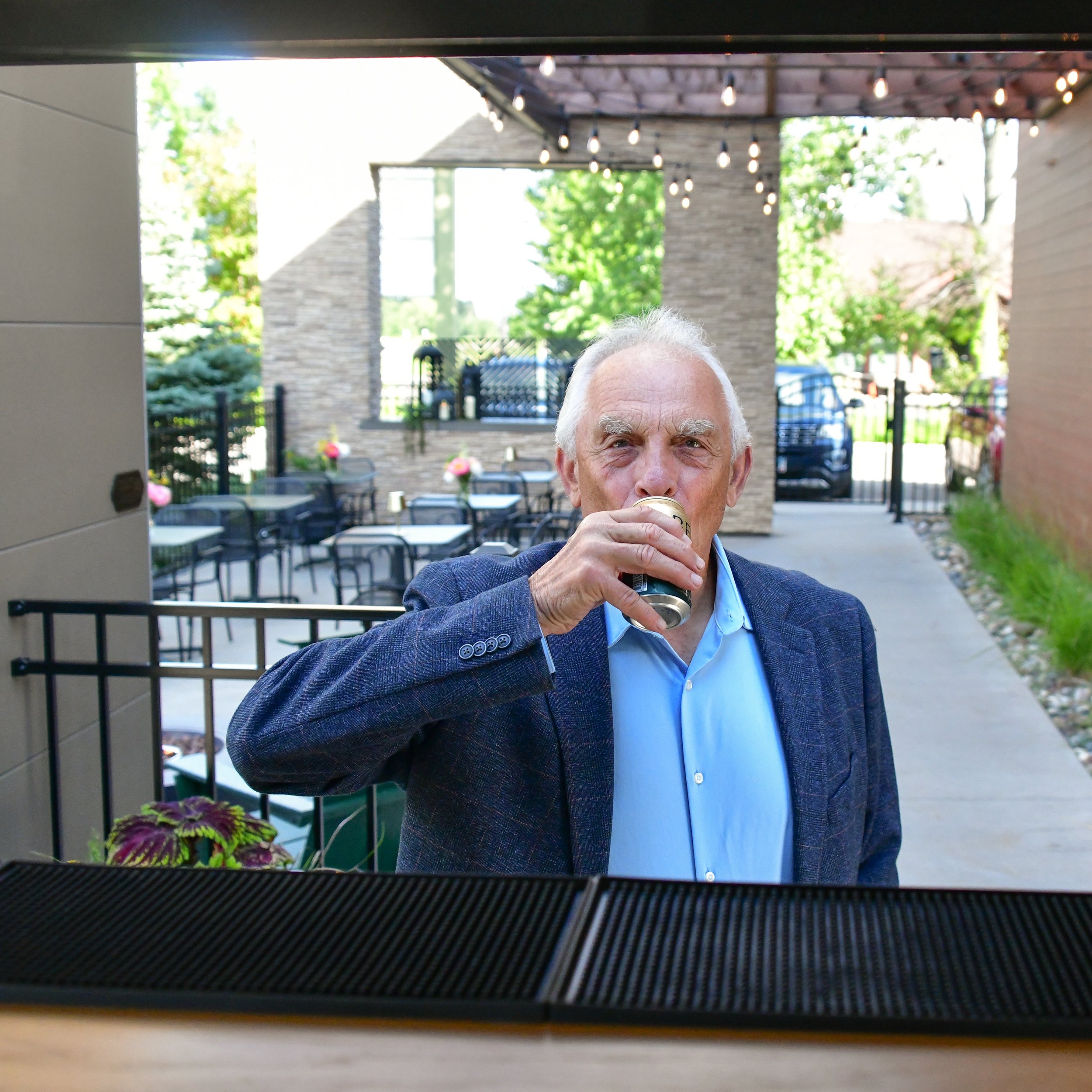 An elderly man with white hair and a blue shirt drinking from a can, viewed from underneath a structure at an outdoor patio area with tables, chairs, string lights, and greenery in the background.