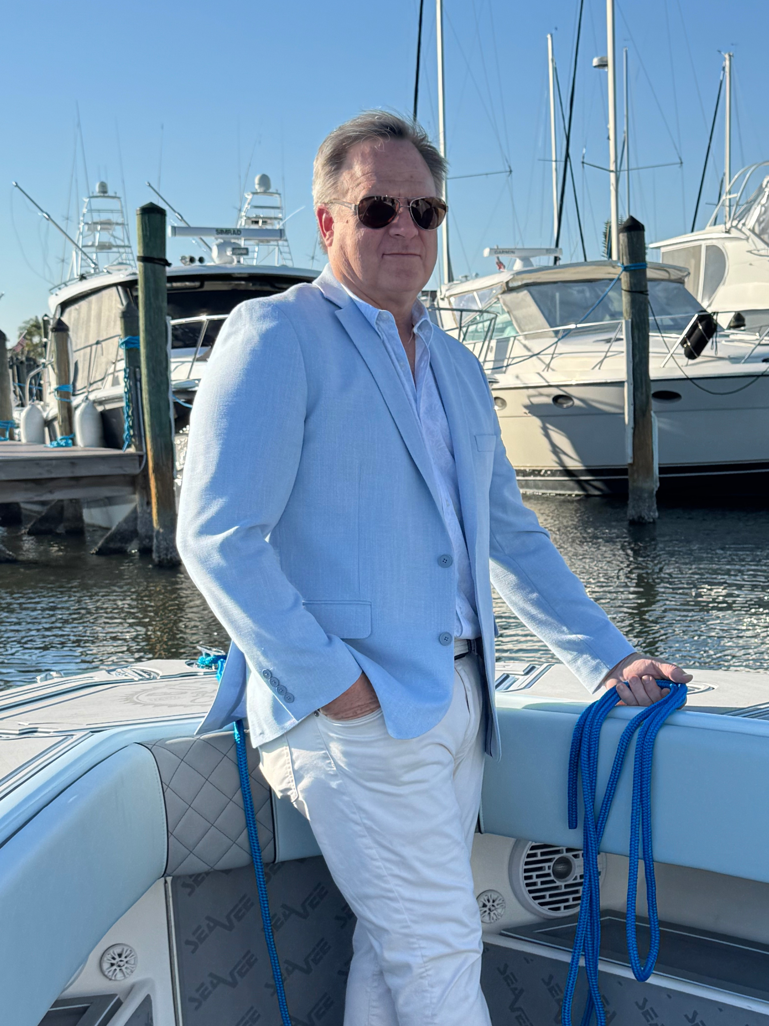 Mark Bouchard standing on the bow of a boat in a Northeast Florida marina, wearing a light blue blazer and sunglasses, with yachts and masts in the background.