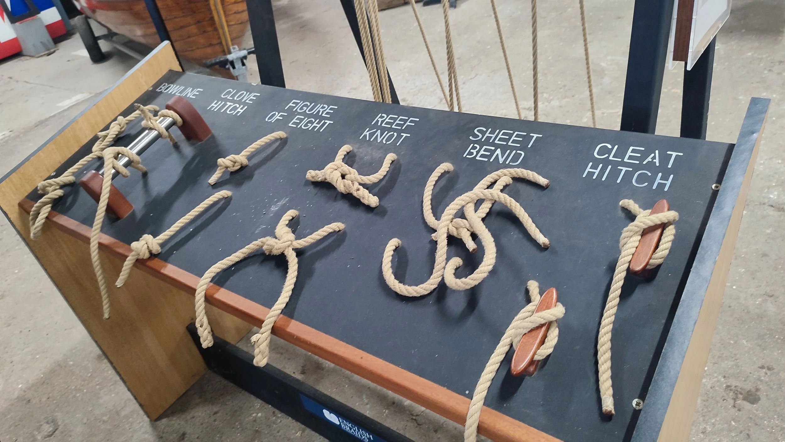 Rope tying demonstration board showing different knots labeled as Bowline Hitch, Clove Hitch, Figure of Eight, Reef Knot, Sheet Bend, and Cleat Hitch, with ropes tied to each knot.