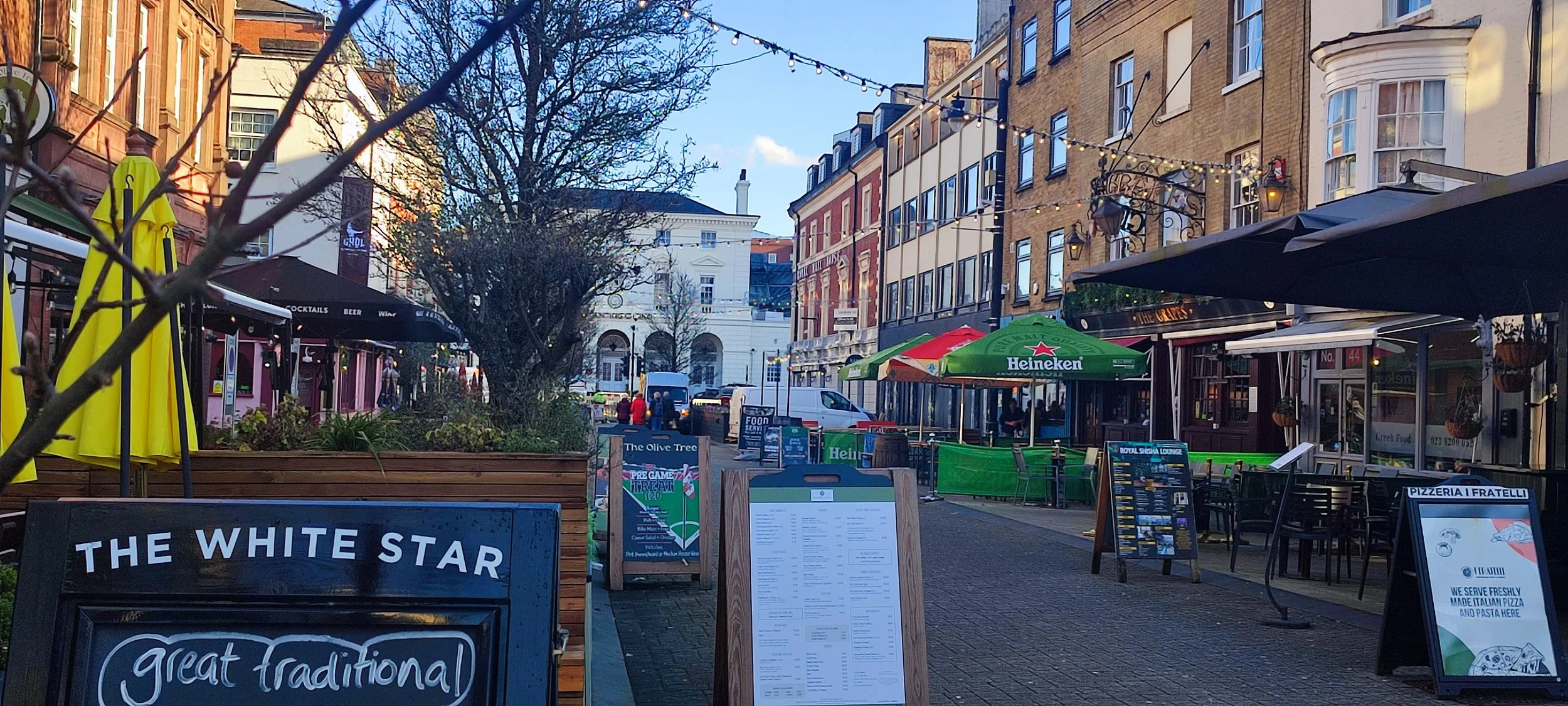 Outdoor street scene with restaurants and cafes featuring tables, chairs, umbrellas, and chalkboards displaying menus, under string lights and a large tree, with historic buildings and a blue sky in the background.