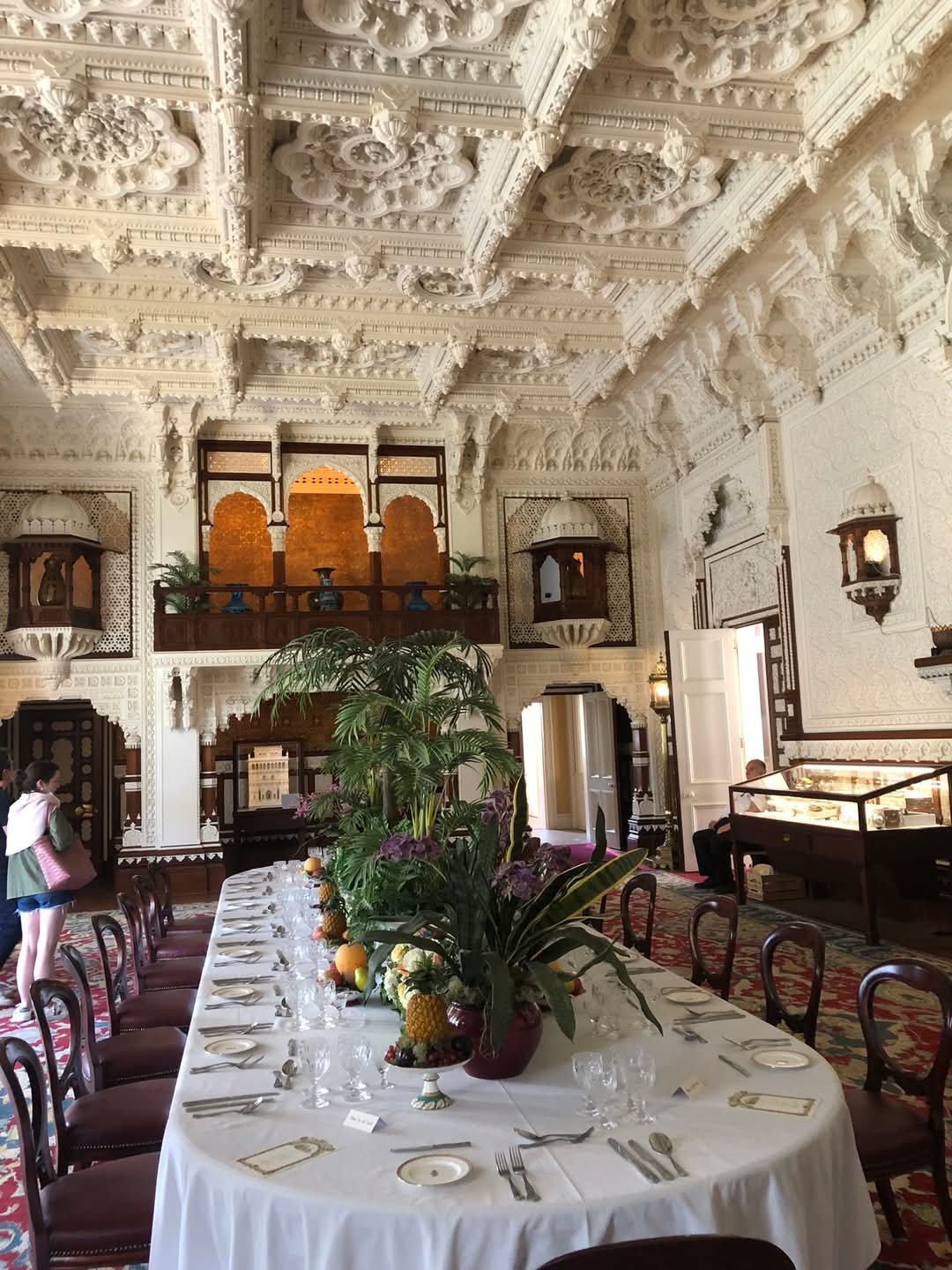 Elegant dining room with ornate white ceiling, long table set with glassware and cutlery, floral centerpiece with greenery and pineapples, large window and intricate wall details.