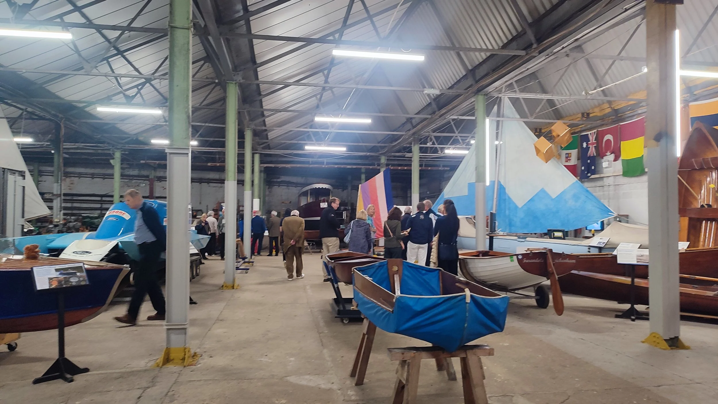 Indoor boat exhibition with various boats on display, including small sailboats and rowboats, and visitors walking around exploring the boats.