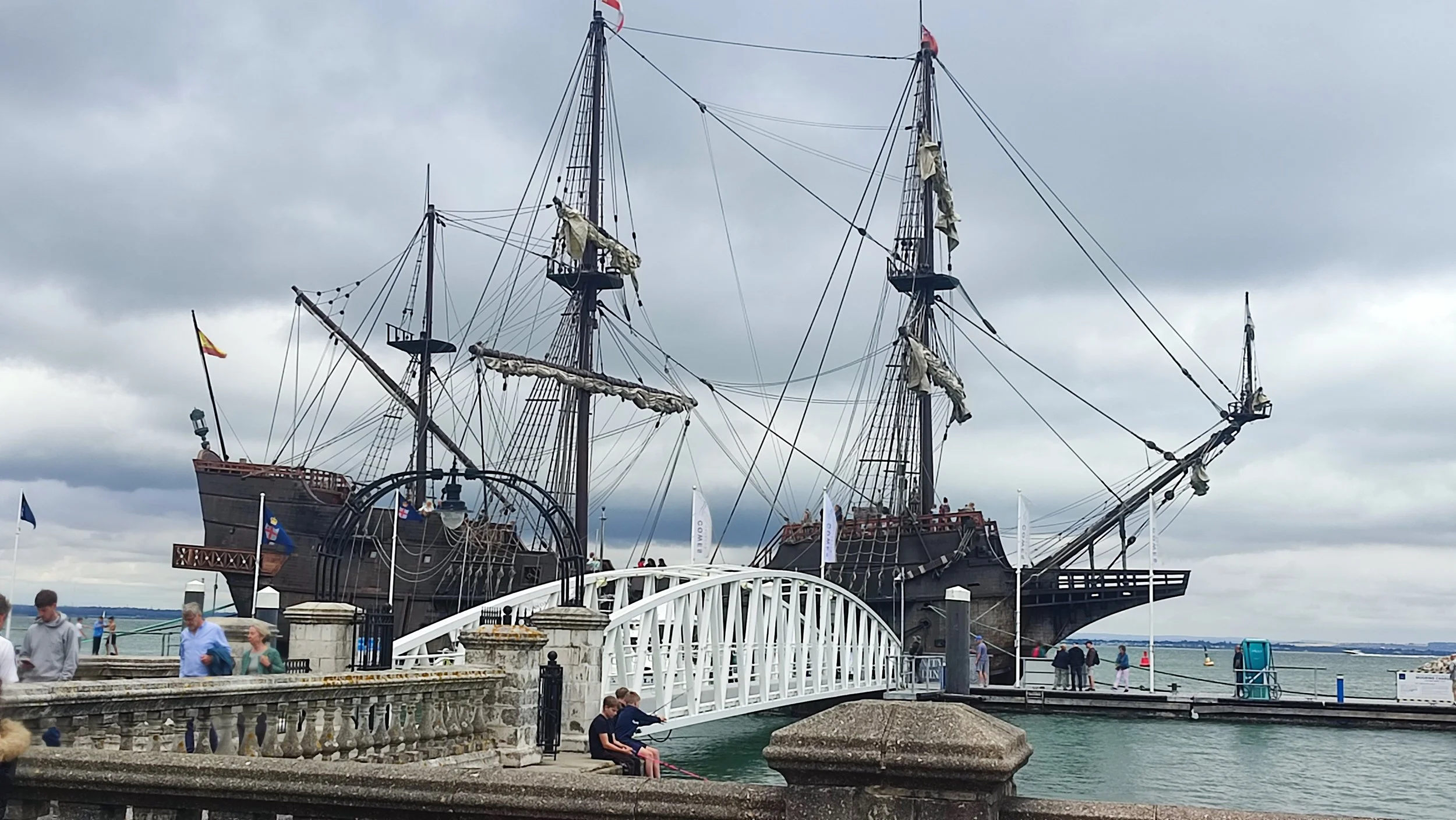 A large, historic pirate ship with multiple masts docked at a waterfront. The ship's black hull and intricate rigging are visible, with some sails furled. In the foreground, there is a small pedestrian bridge and several people walking or sitting near the water, under a cloudy sky.