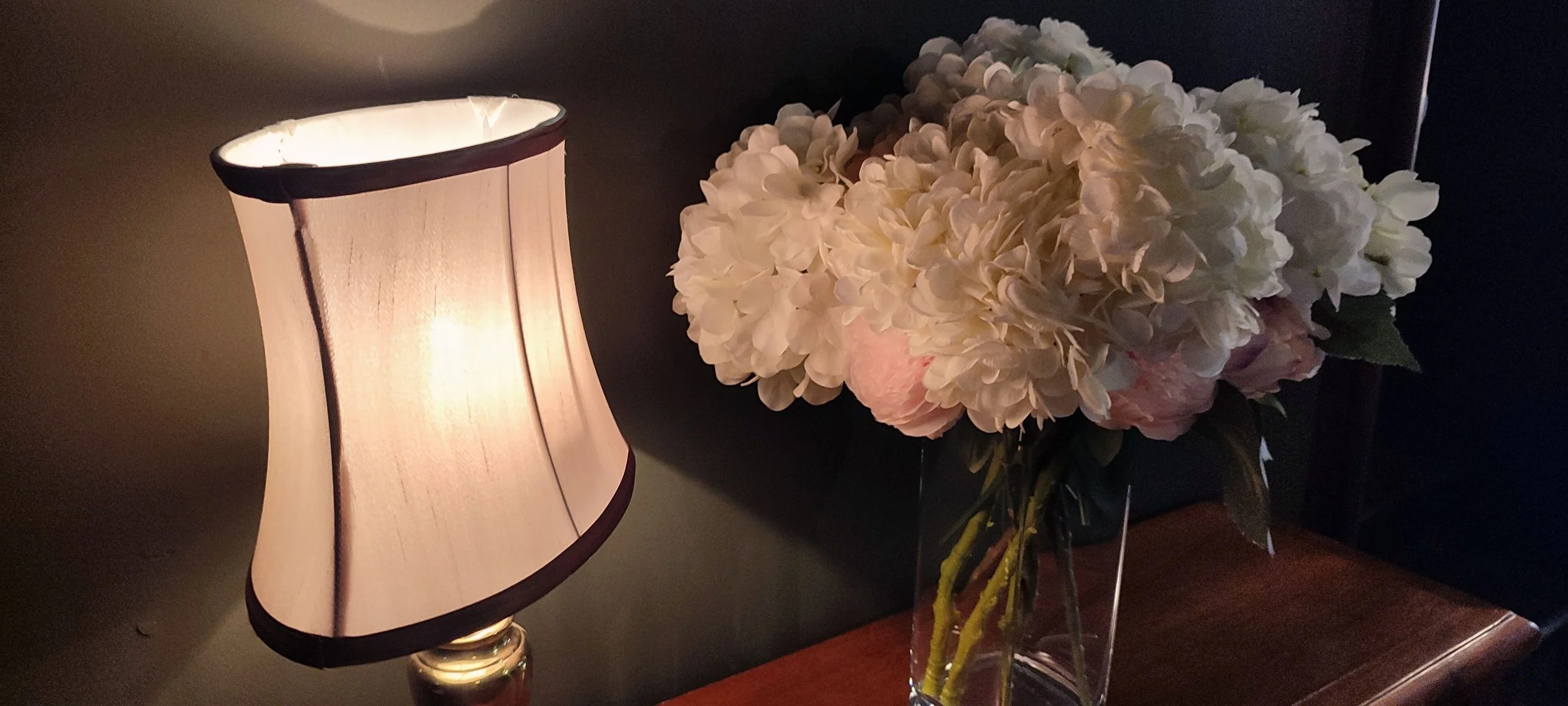 A soft glowing table lamp beside a large, white hydrangea flower arrangement in a clear glass vase on a wooden surface.