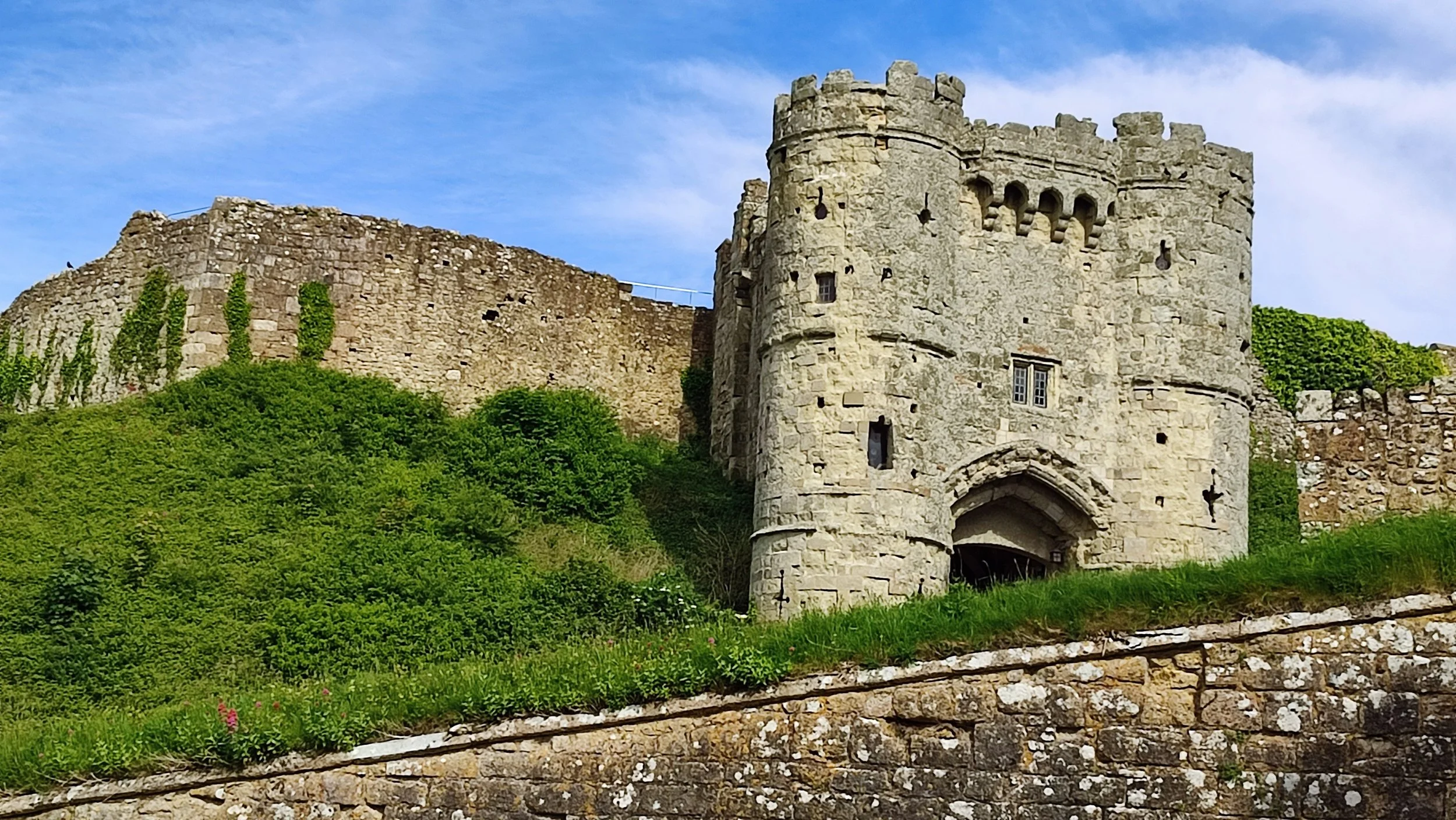 A historic stone castle with a round tower, stone walls, and crenellations, surrounded by green grass and bushes under a partly cloudy blue sky.