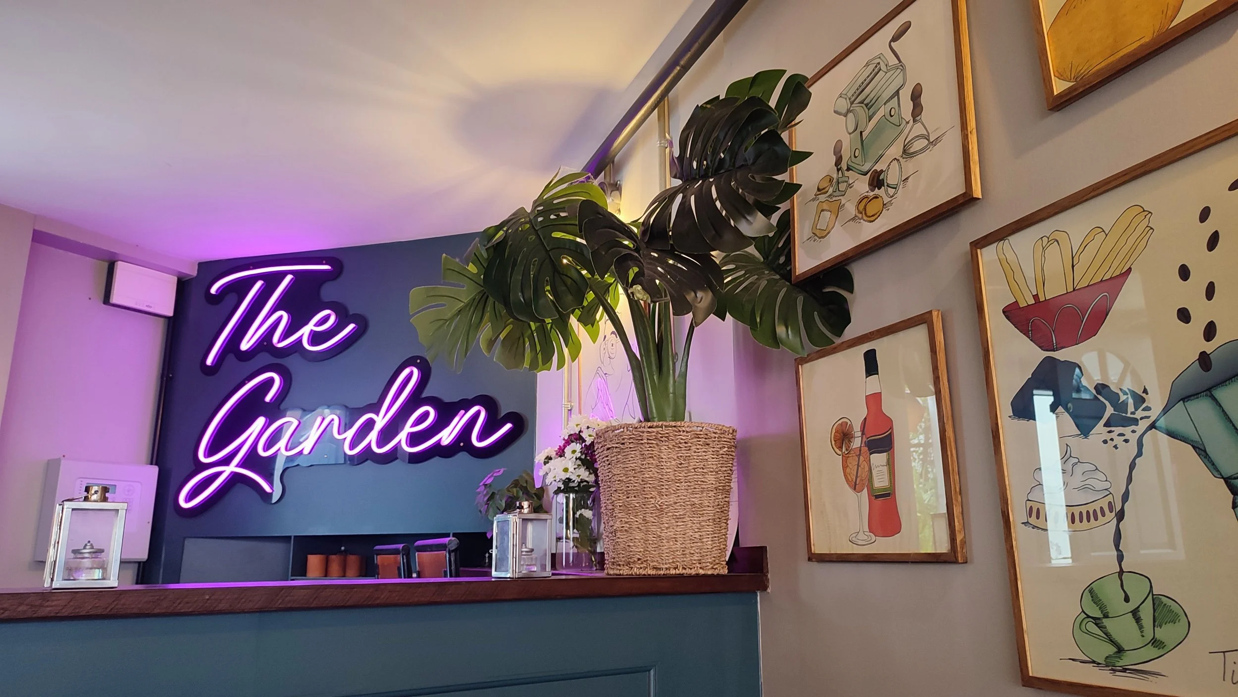 Interior of a cafe with a purple neon sign that says 'The Garden,' a large potted Monstera plant, framed artwork of food and drinks on the wall, and a wooden counter.