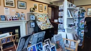 Interior of a bookstore or gift shop with shelves of postcards, photographs, and framed pictures, and a white display rack filled with greeting cards or postcards.