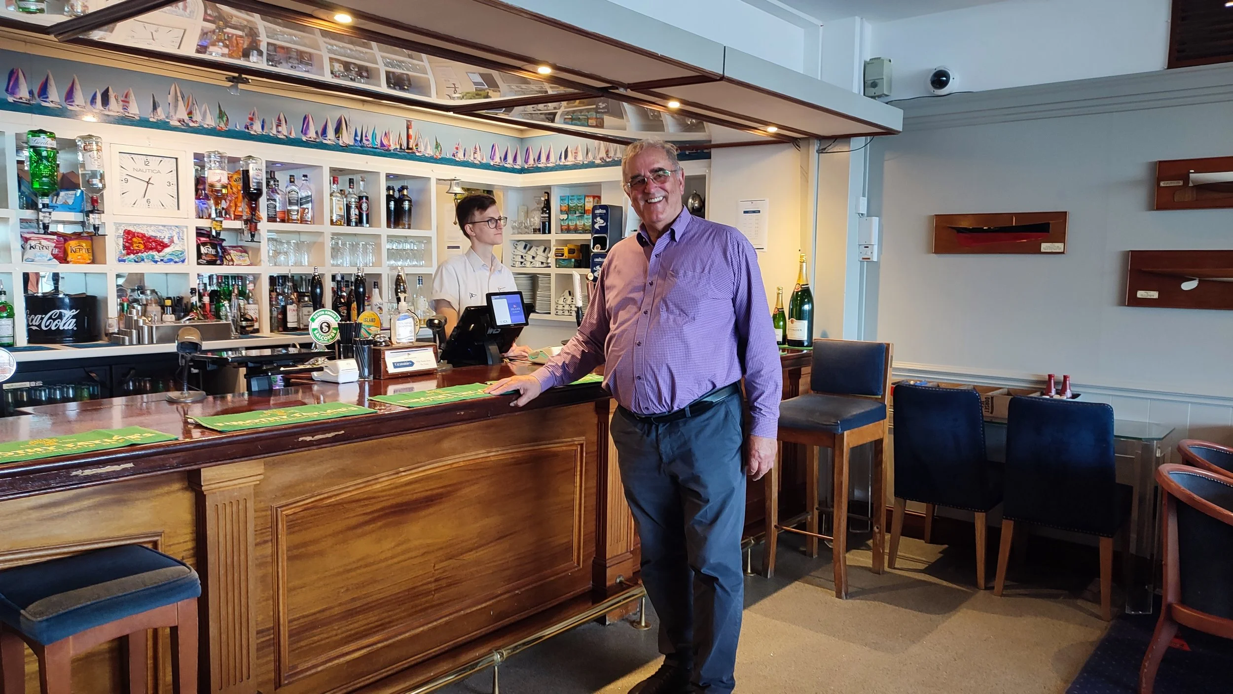 A man in a purple shirt and dark pants smiling at the camera standing at a bar in a restaurant or pub. Behind the bar are shelves with bottles, glasses, and decorations, with a bartender and another person working behind the counter. The setting appears to be a casual dining or drinking establishment.