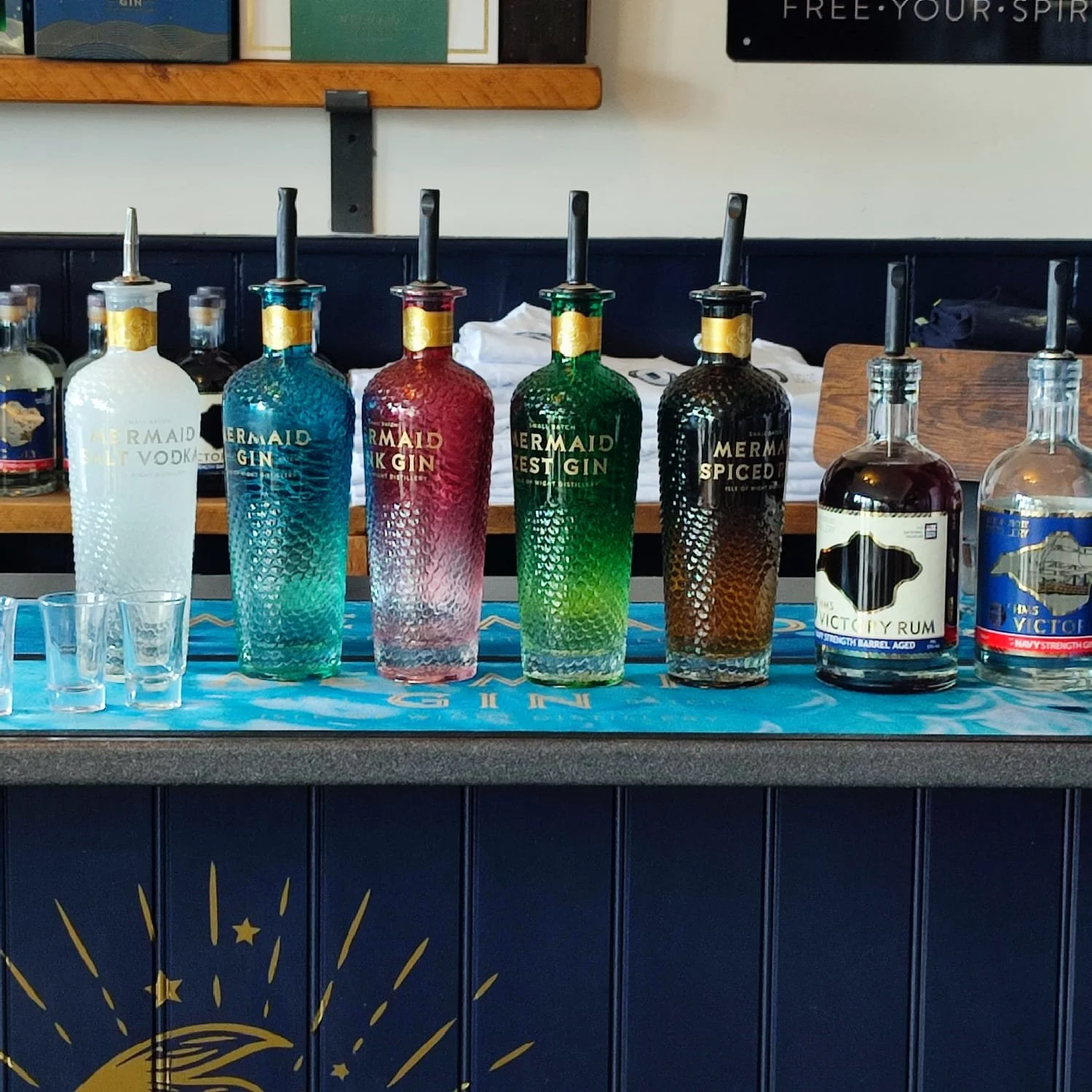 A display of six colorful bottles of spirits with spouts on a bar counter, labeled from left to right: Mermaid Salt Vodka, Mermaid Gin, Mermaid Dark Gin, Mermaid Zest Gin, Mermaid Spiced Rum, Victory Rum, with small shot glasses in front of them.
