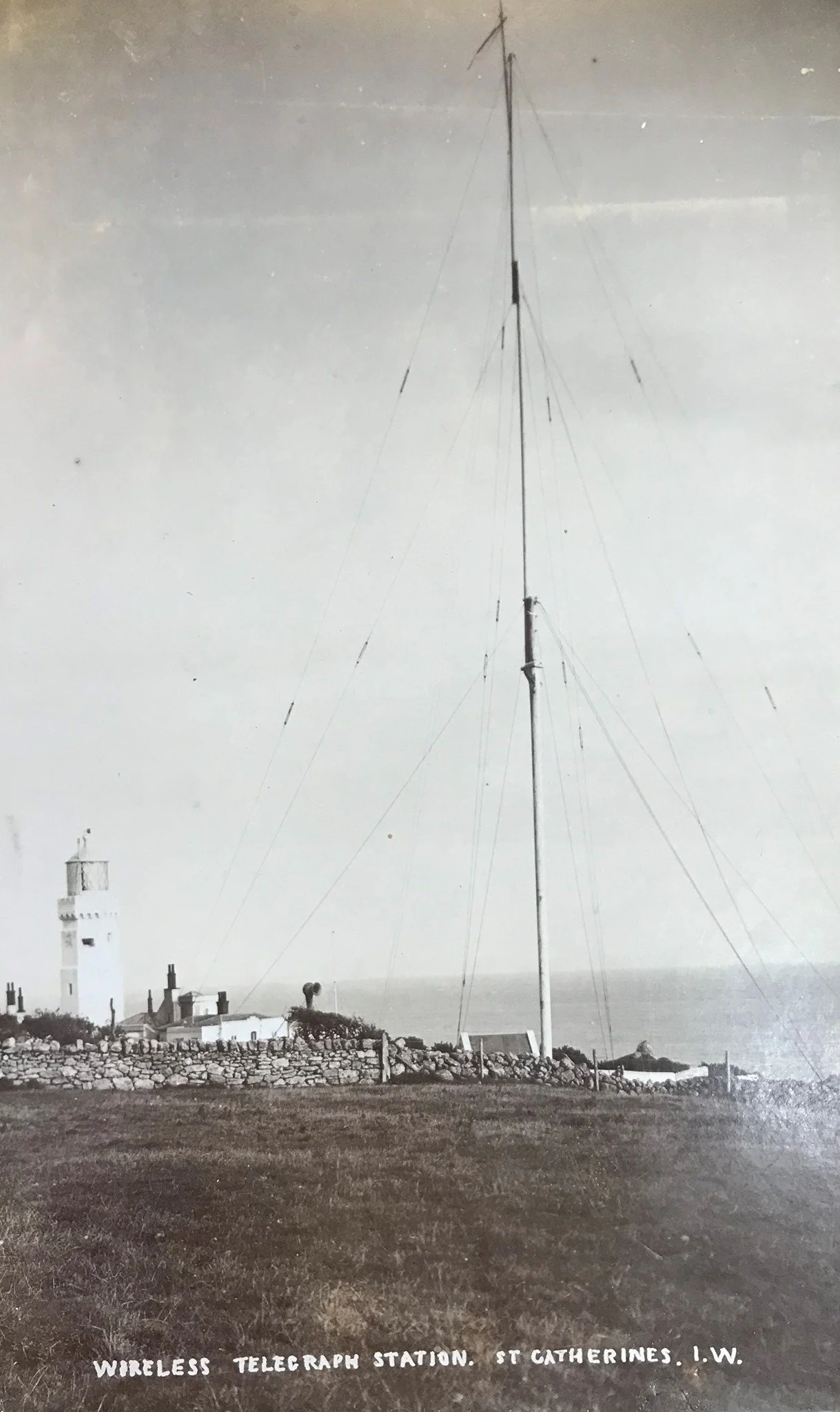 A black and white photograph of a wireless telegraph station at St. Catherines, L.W. with a tall antenna and a lighthouse in the background.