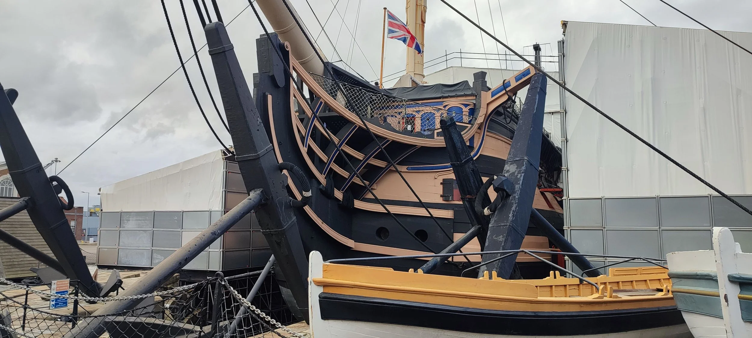 A large historic sailing ship, held upright on supports, with a Union Jack flag on the mast, surrounded by fencing and buildings in the background.