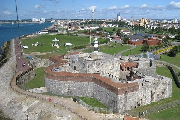 A historic fortress with stone walls and towers overlooking a harbor, with a city skyline in the background, featuring modern buildings and a bridge.