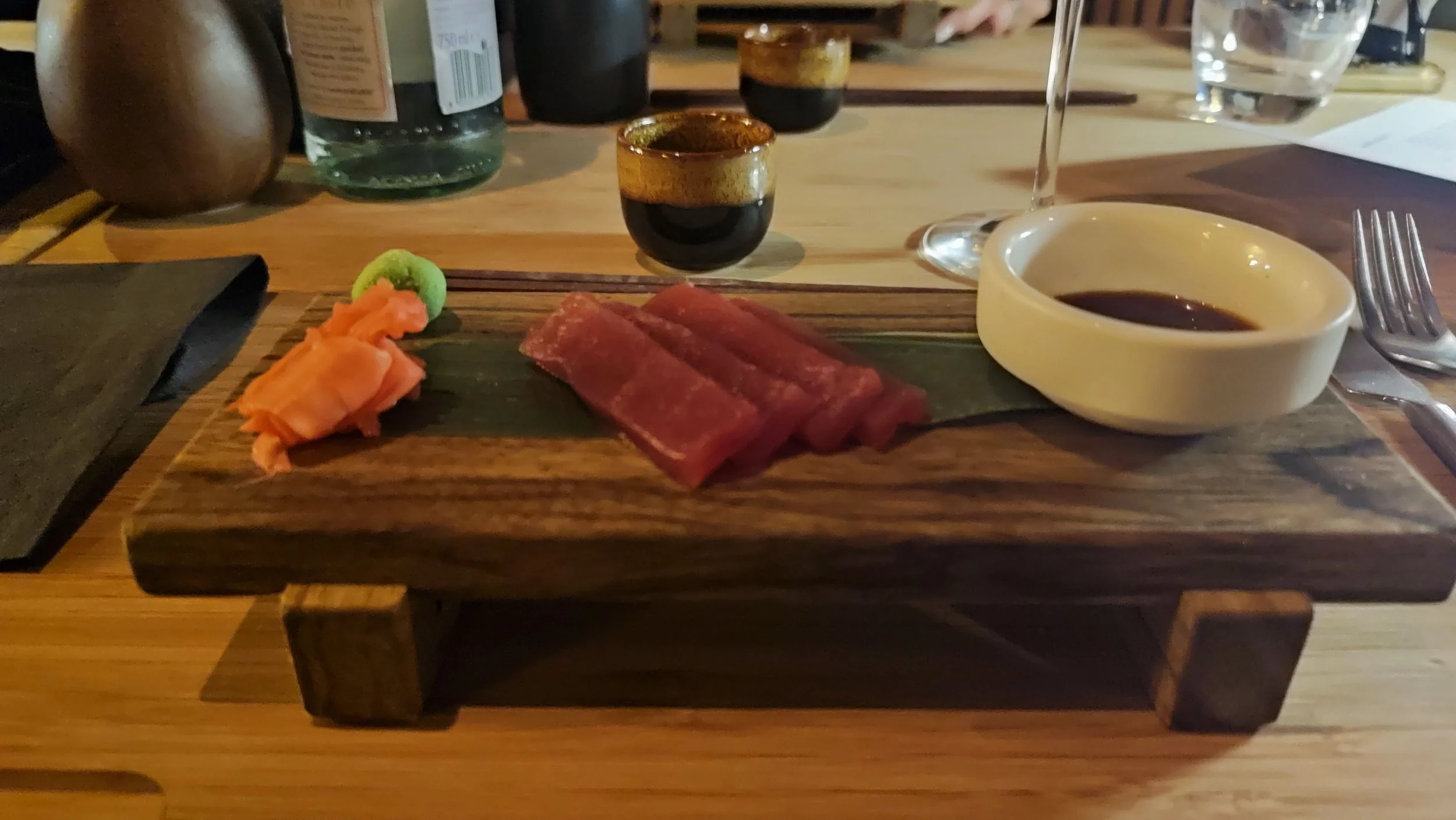 A wooden serving board with slices of raw tuna, a small serving of pickled ginger, wasabi, and a bowl of dipping sauce, with two small cups of soy sauce in the background on a restaurant table.