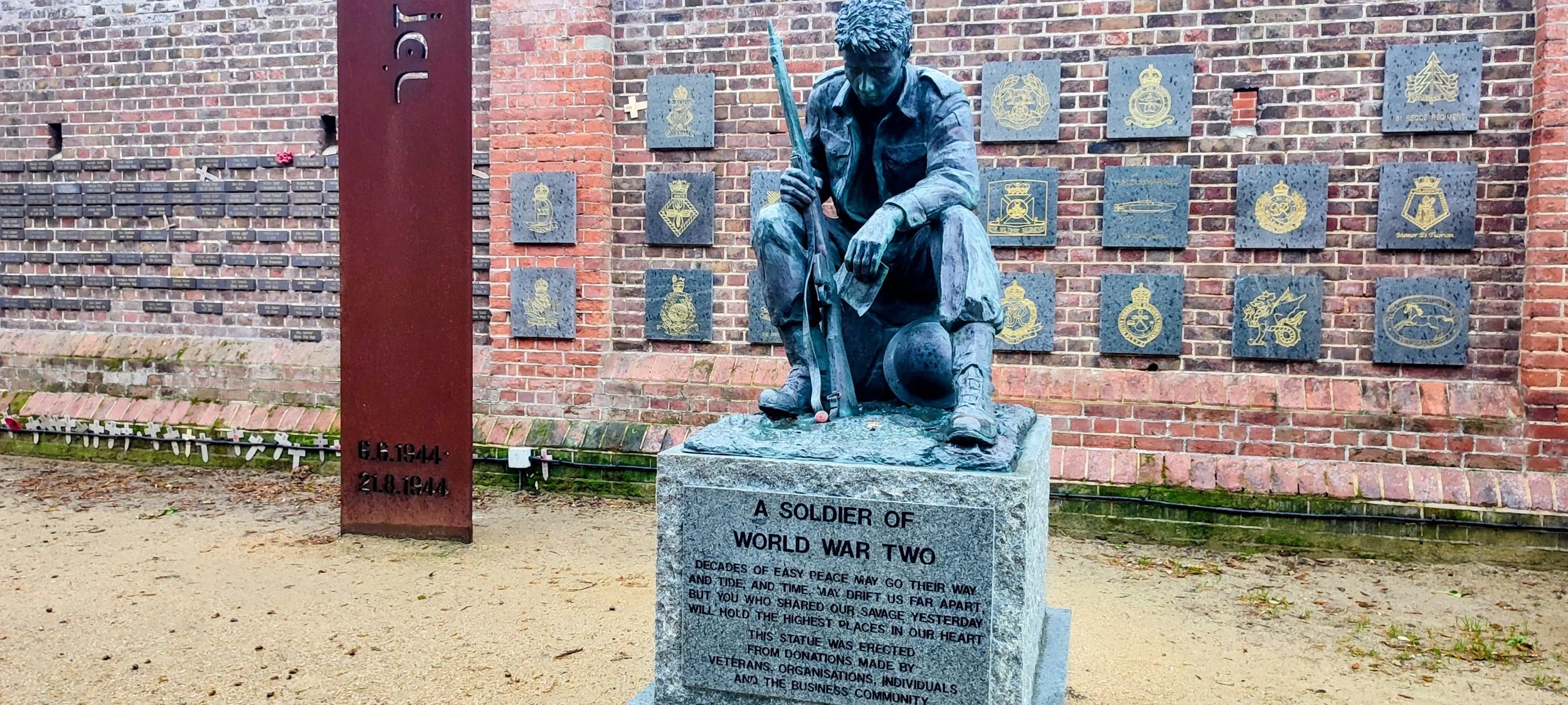 Bronze statue of a World War II soldier sitting with a rifle and helmet, mounted on a stone pedestal with an inscription. Behind the statue, there is a brick wall adorned with plaques and insignias. To the left, a rusty metal post with dates 6.6.1944 and 21.8.1944 engraved.