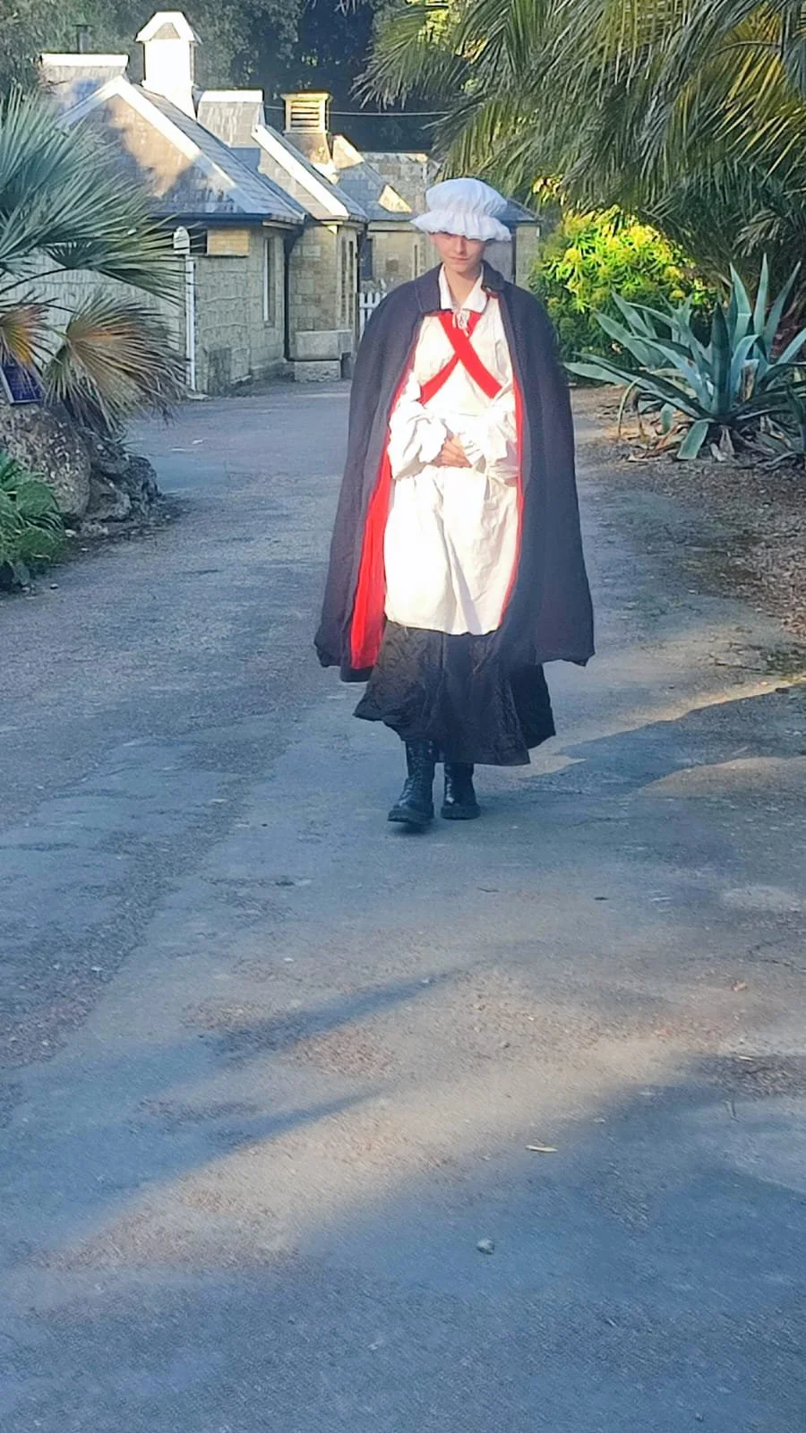 Person dressed in historical costume walking on a dirt path with plants and houses in the background.