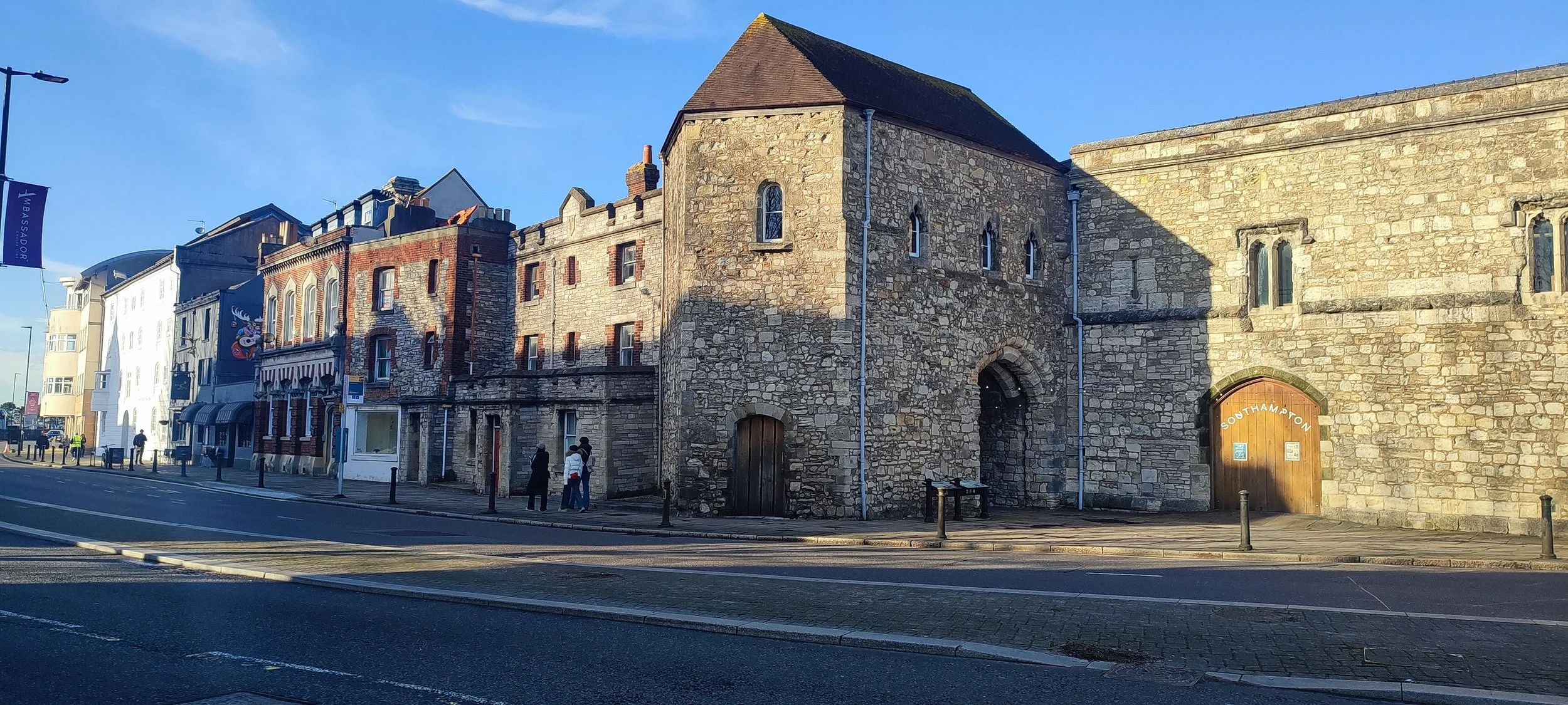 Street view of historic stone and brick buildings, including a church with a wooden door labeled 'Southampton,' with pedestrians walking and a clear blue sky.
