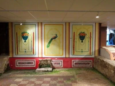 Three framed artworks on a wall: two decorative vases with flowers and a peacock in the center, with a red and pink base wall and a pile of rocks or bricks on the floor.