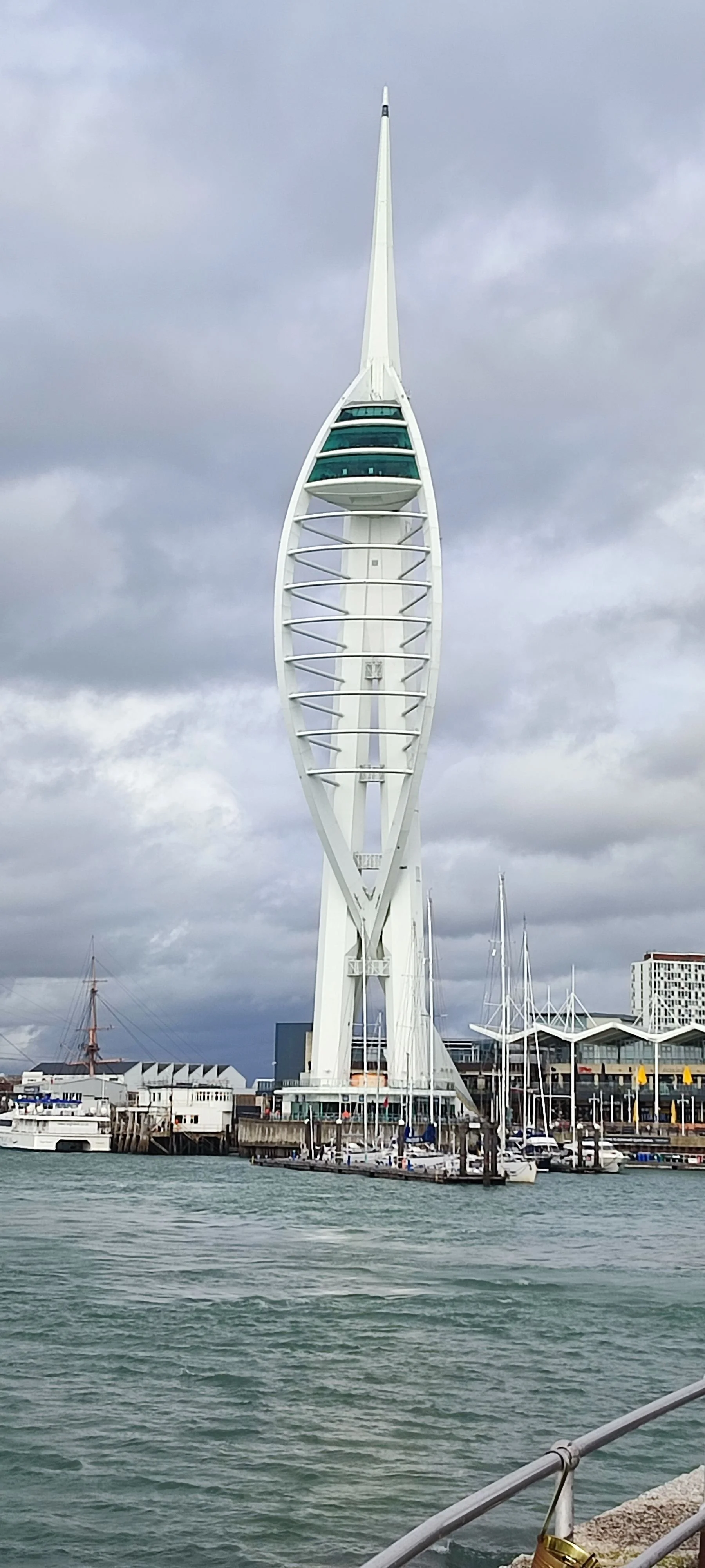 Sydney Tower Eye, a tall, modern observation tower with a distinctive design, skyline, water, boats docked at the pier, and a cloudy sky.
