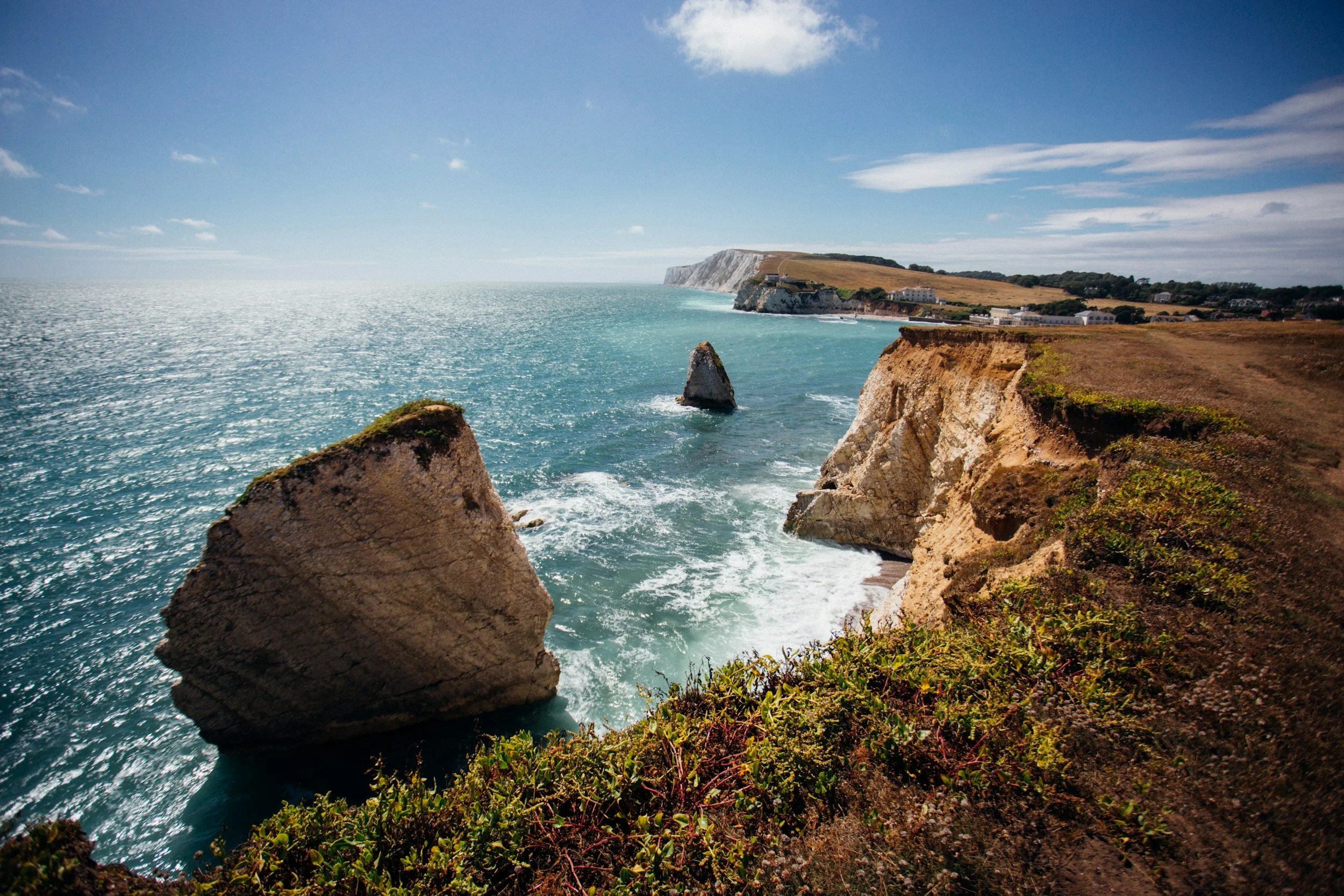Cliffs along a coastline with large rocks in the water and a village in the distance under a blue sky with clouds.