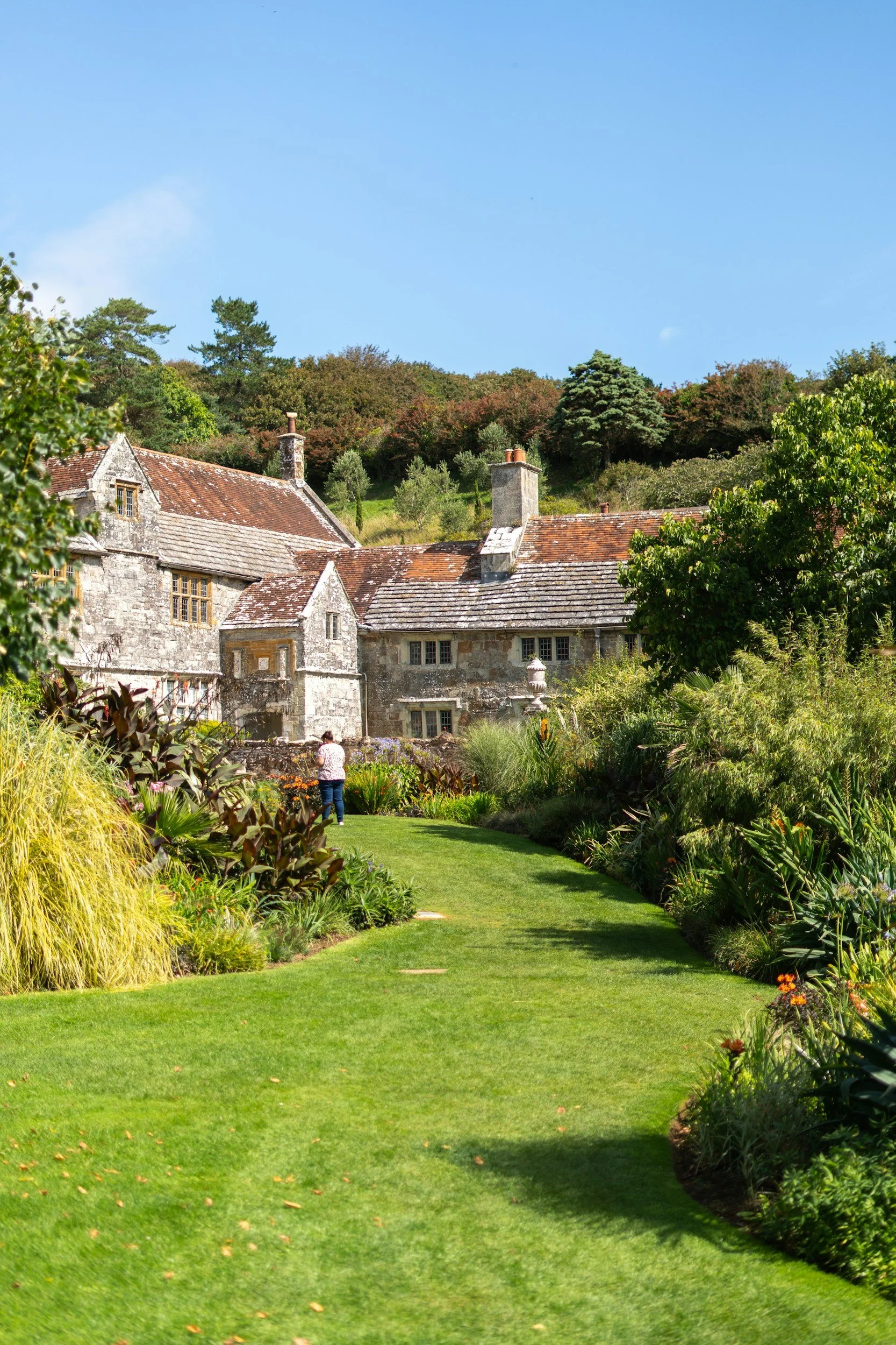 A scenic view of a well-maintained garden with green grass, bushes, and trees leading up to an old stone house with multiple chimneys, set against a backdrop of a hill with more trees, under a clear blue sky.