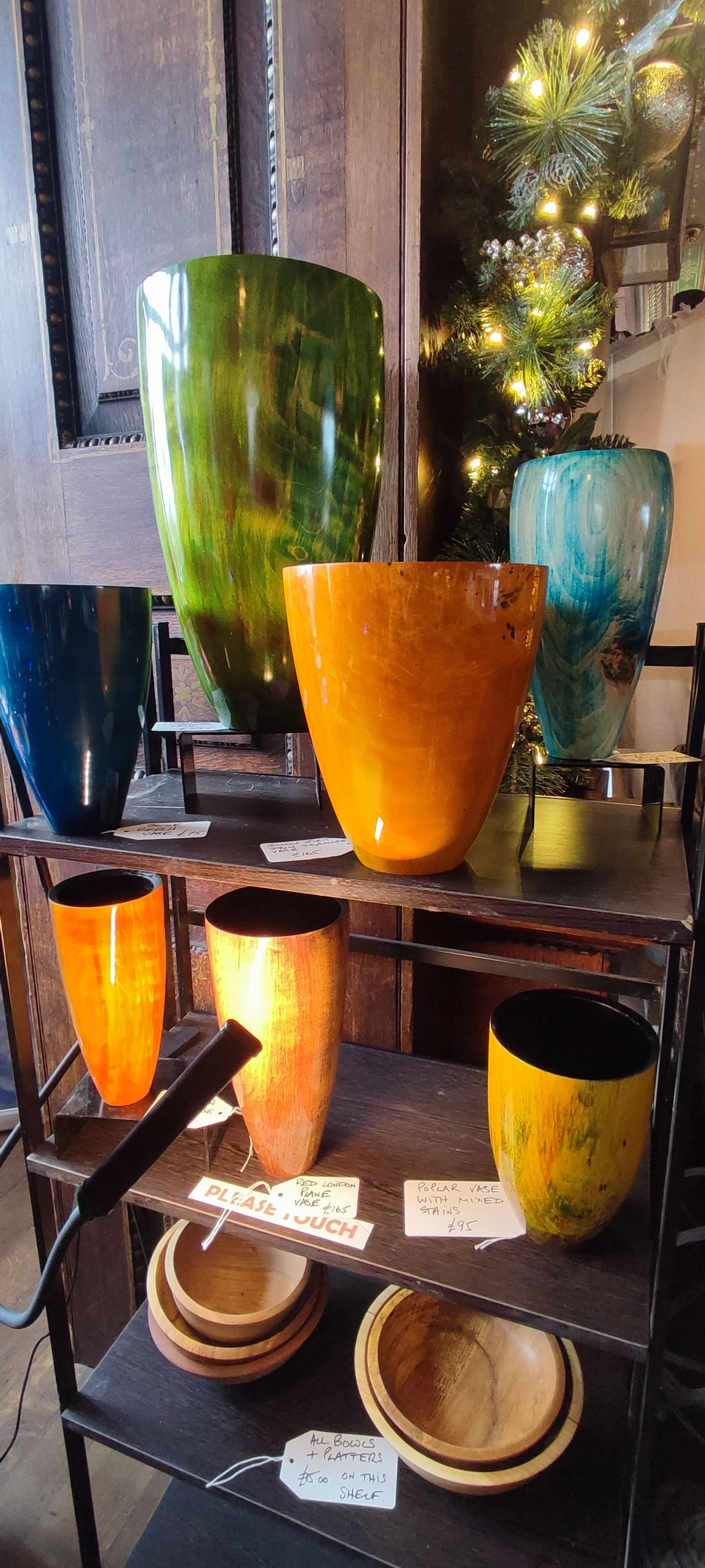 Display of colorful glass vases on a dark wooden shelving unit with wooden bowls underneath, decorated with holiday lights and a Christmas tree in the background.