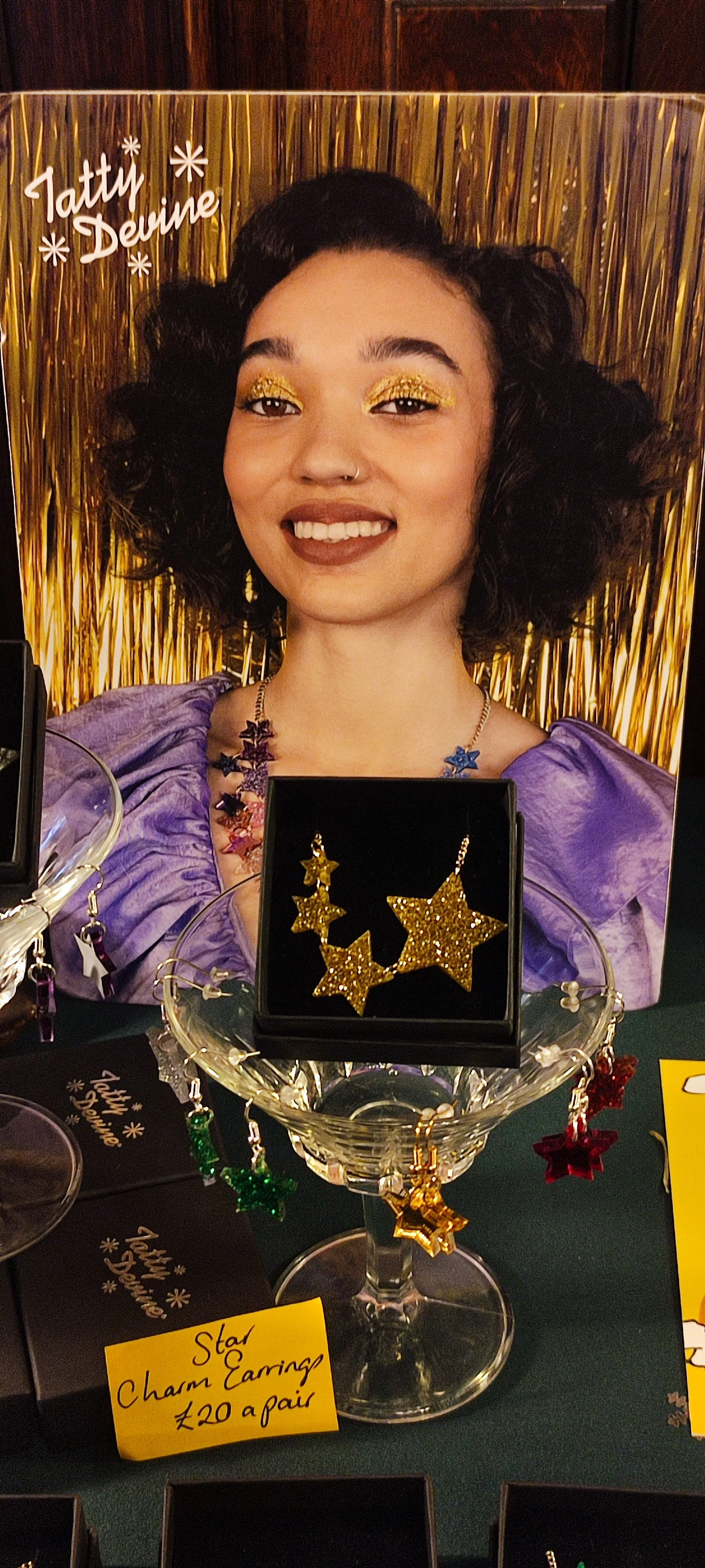 A display at a booth showcasing gold glitter star-shaped earrings with a sign saying 'Star Charm Earrings £20 a pair'. The backdrop features a large photo of a smiling woman with dark curly hair, gold eyeshadow, a nose ring, and a purple top.