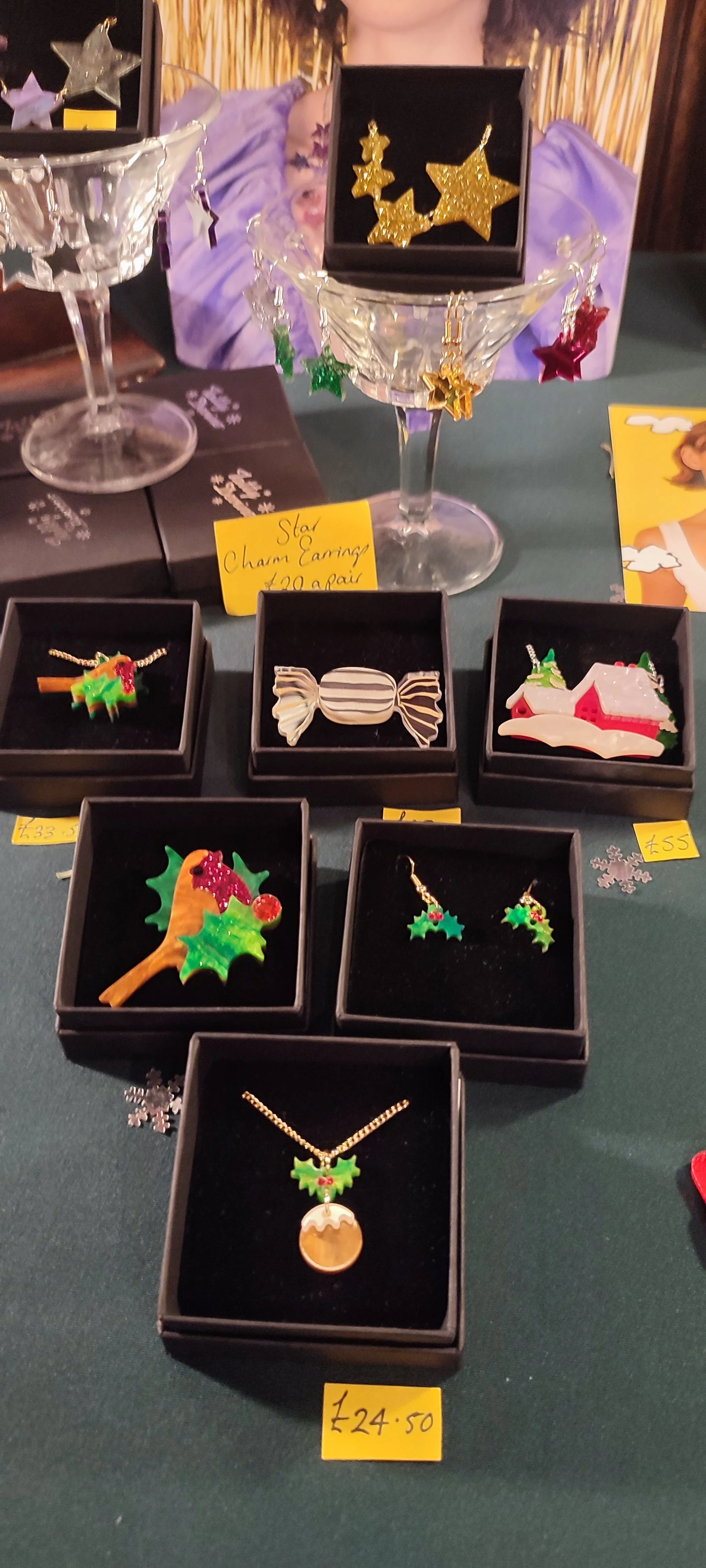 Holiday-themed jewelry including earrings and necklaces inside black display boxes on a table, with Christmas and festive decorations.