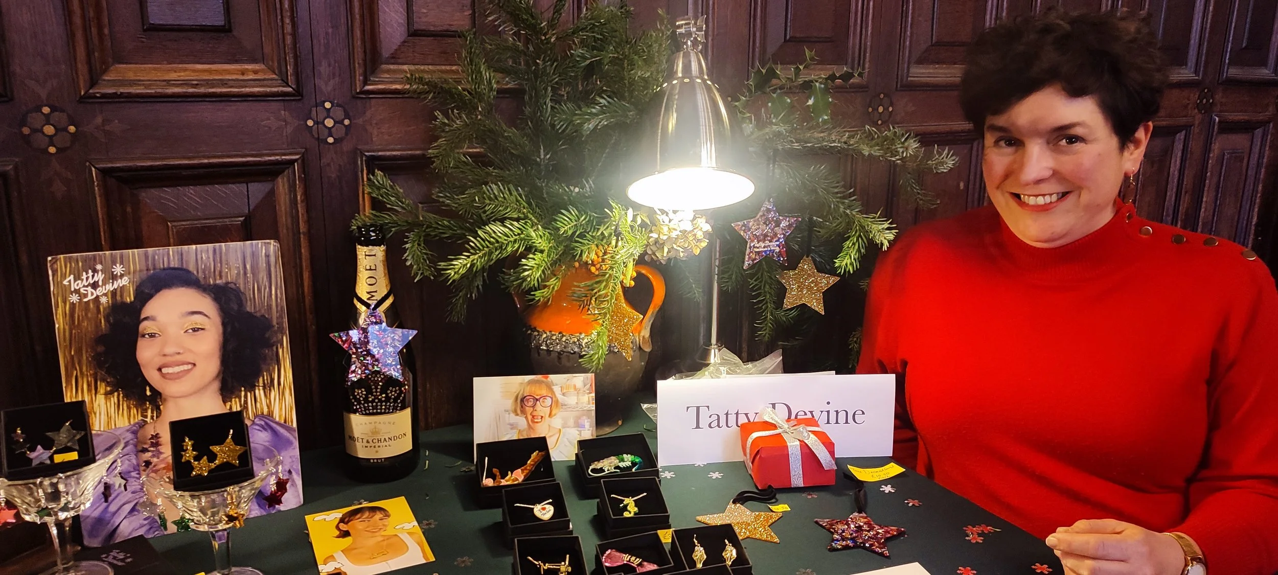 A woman with short dark hair and a big smile, wearing a red sweater with button details on the shoulder, sitting at a festive table with Christmas decorations, jewelry display, and a photo of a woman named Tatty Devine, in a room with dark wooden wall panels and an illuminated lamp.