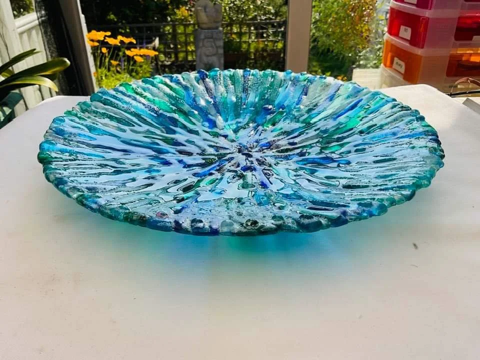 A large, round, textured glass bowl with a blue and green swirled pattern on a white surface outdoors, with a garden and railing in the background.