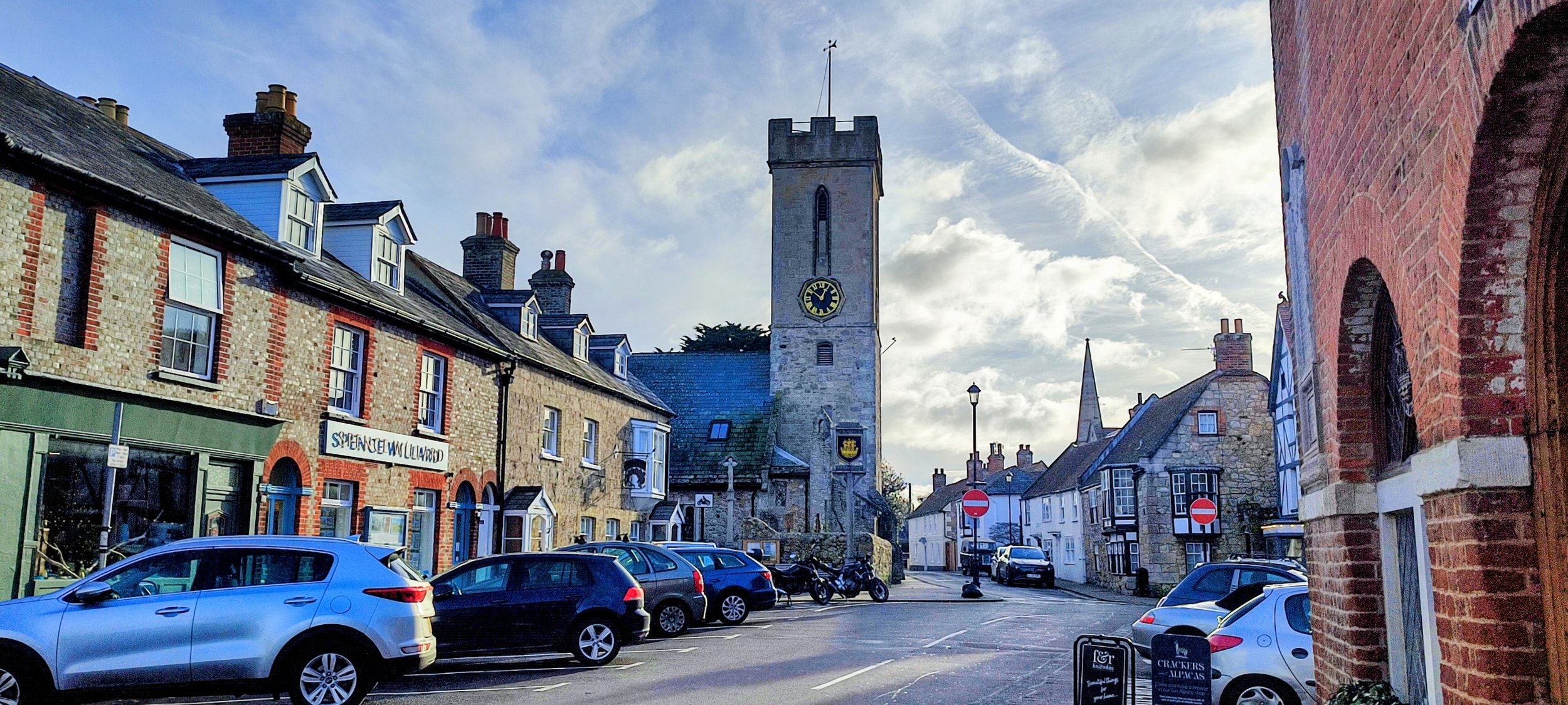 A street view in a small town with cars parked along the road. There are brick buildings on both sides, and a clock tower in the background under a partly cloudy sky.