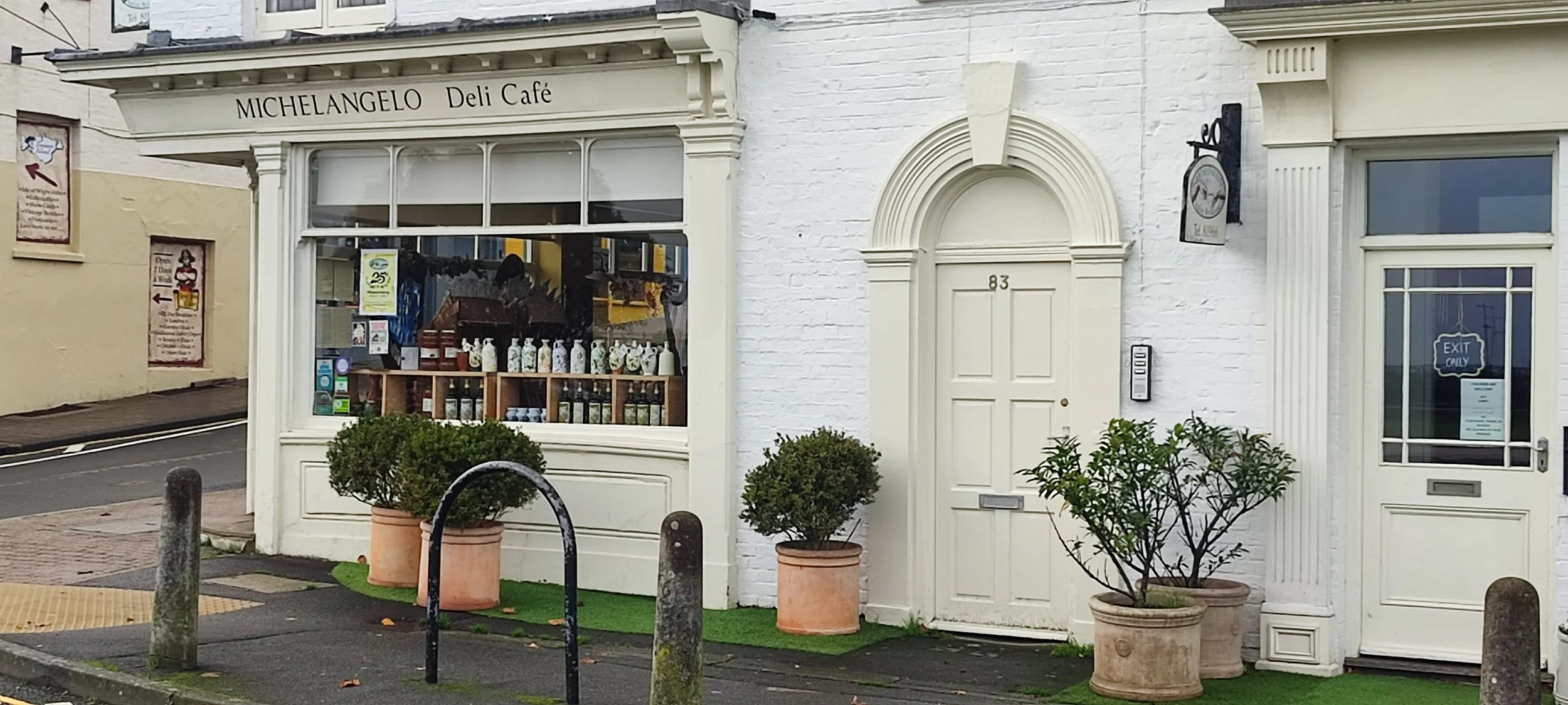 Exterior of Michelangelo Deli Café with a white brick building, potted plants, and a large window displaying bottles, with the address 83 on the door.