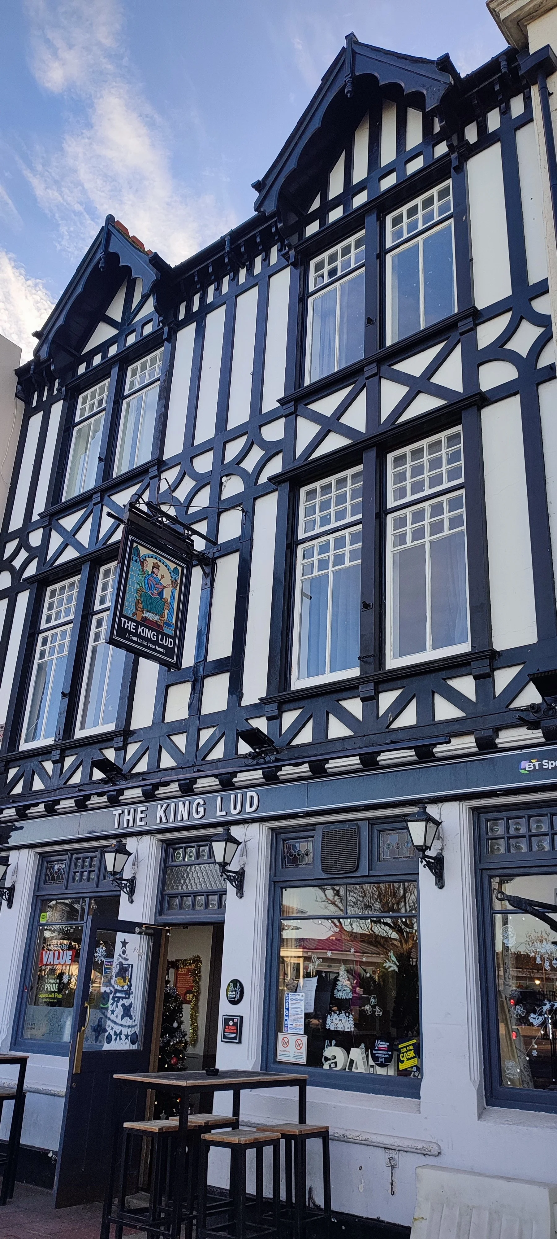 Exterior of a black and white Tudor-style building with the sign "The King Lud" on the facade, decorated for Christmas with a Christmas tree visible through the window.