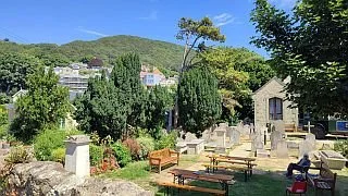 A historic graveyard with old stone tombstones, lush green trees, and a hillside village in the background.