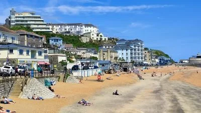 Beachside scene with sandy shore, people sitting and walking, and buildings on a hillside in the background under a blue sky.