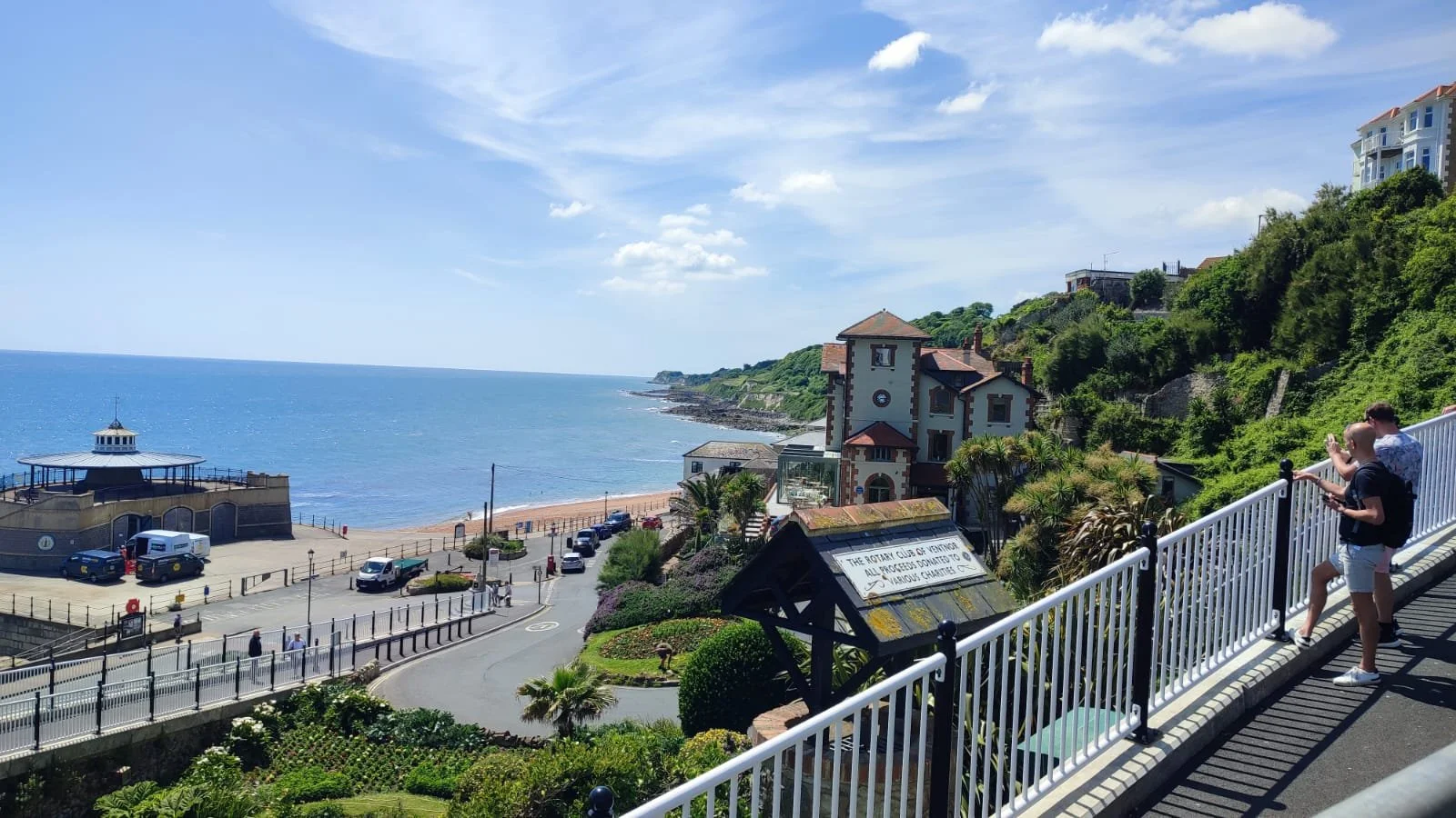 People overlooking a scenic coastal view from a promenade with a white railing, a clock tower, residential buildings, and a road near the beach on a sunny day with blue sky and scattered clouds.