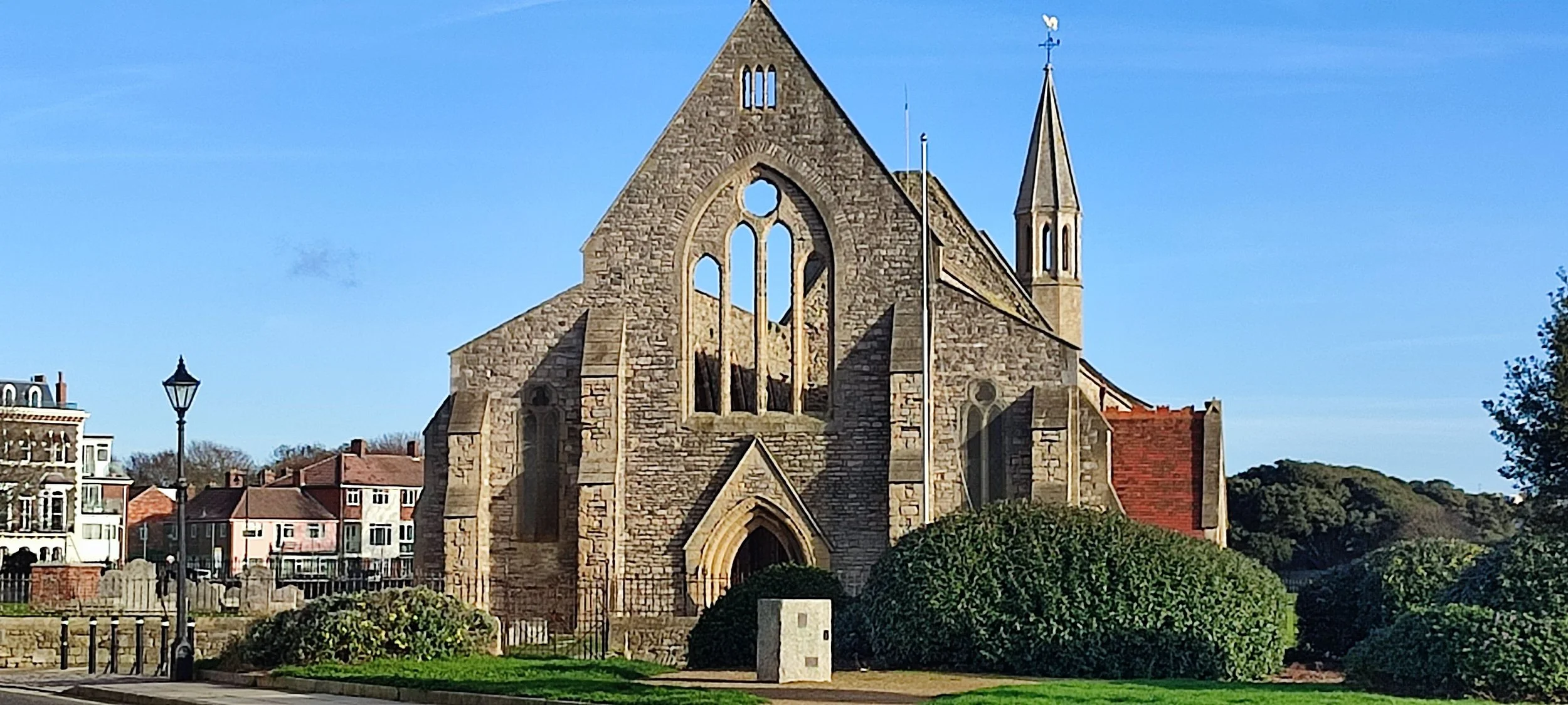 Stone church with large arched windows, small steeple with weather vane, surrounded by green bushes and trees, under a clear blue sky.