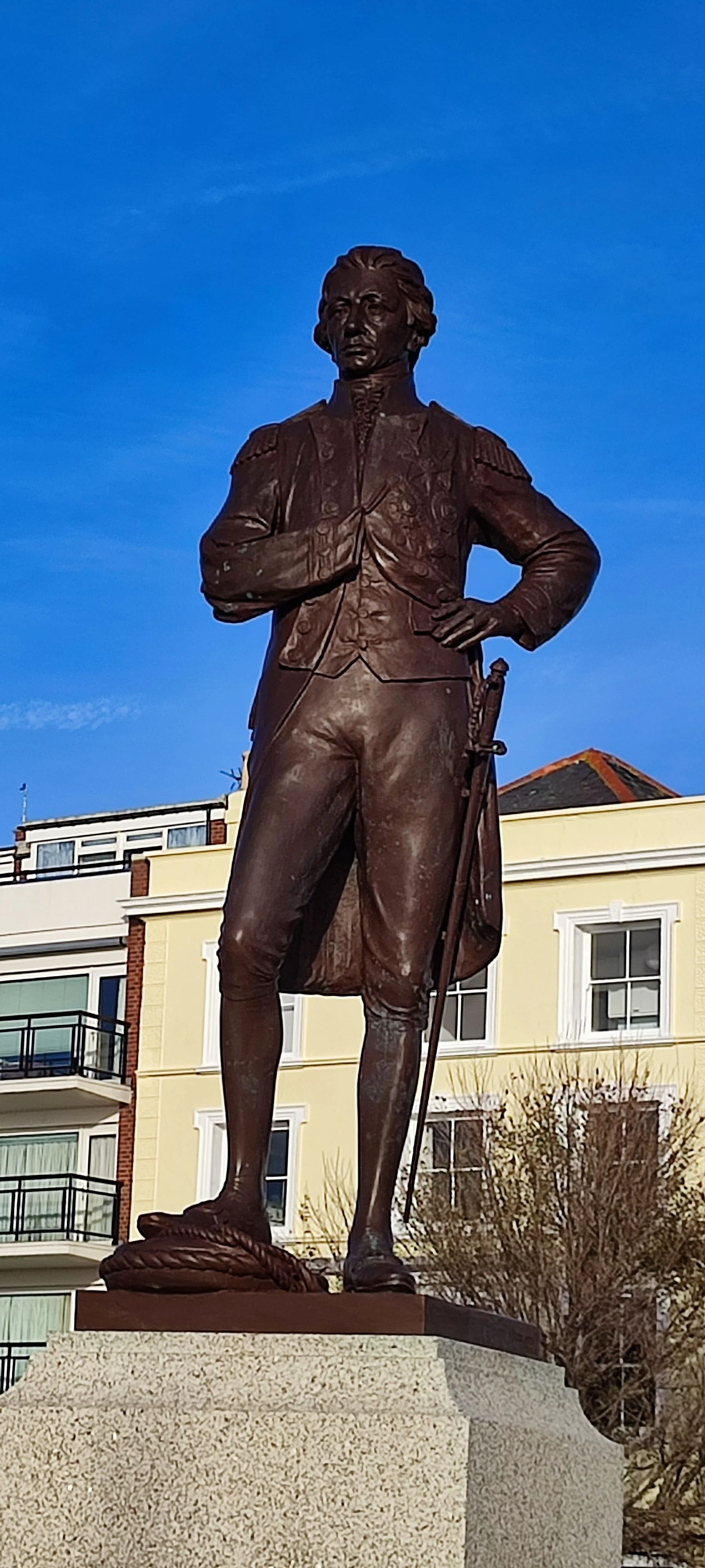 Bronze statue of a man in a military uniform with a sword, standing on a stone pedestal in front of residential buildings.