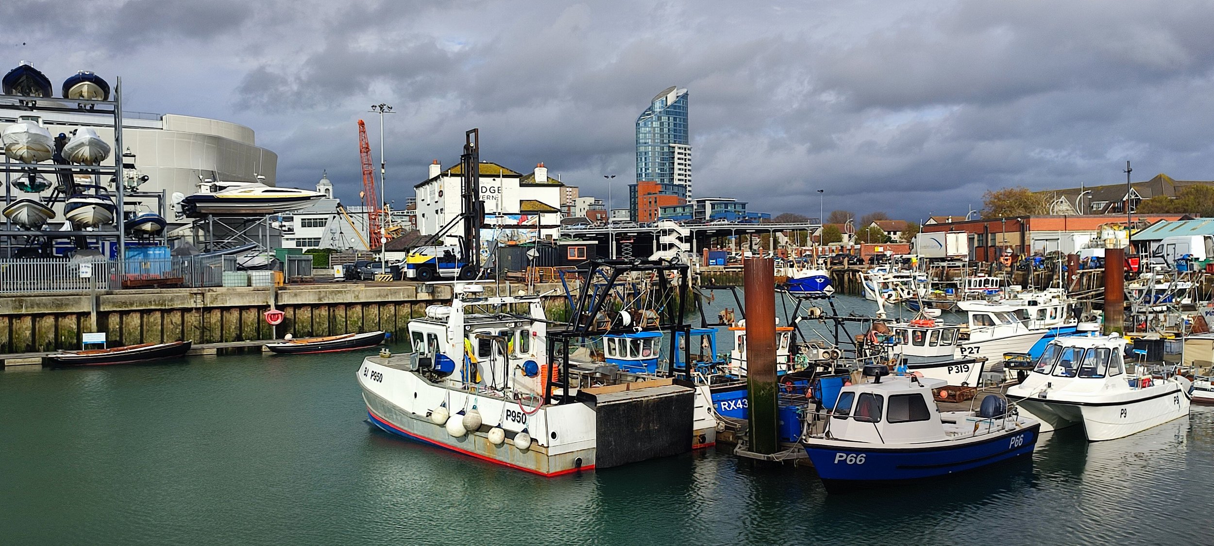 Boats docked in a harbor with city buildings and a cloudy sky in the background.