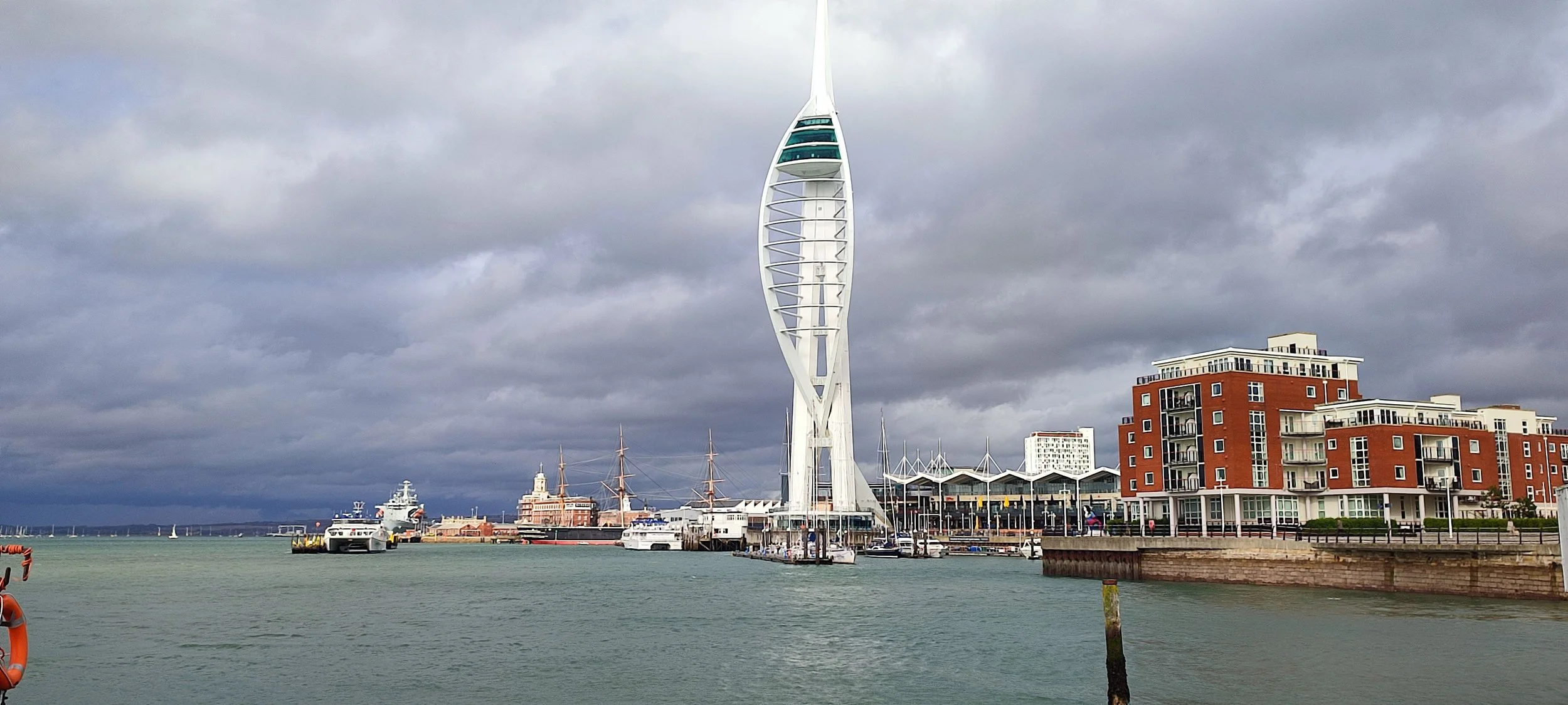 A waterfront scene in Brighton, UK, featuring the British Airways i360 observation tower in the center, with residential buildings to the right, ships and sailboats on the water, and a cloudy sky overhead.