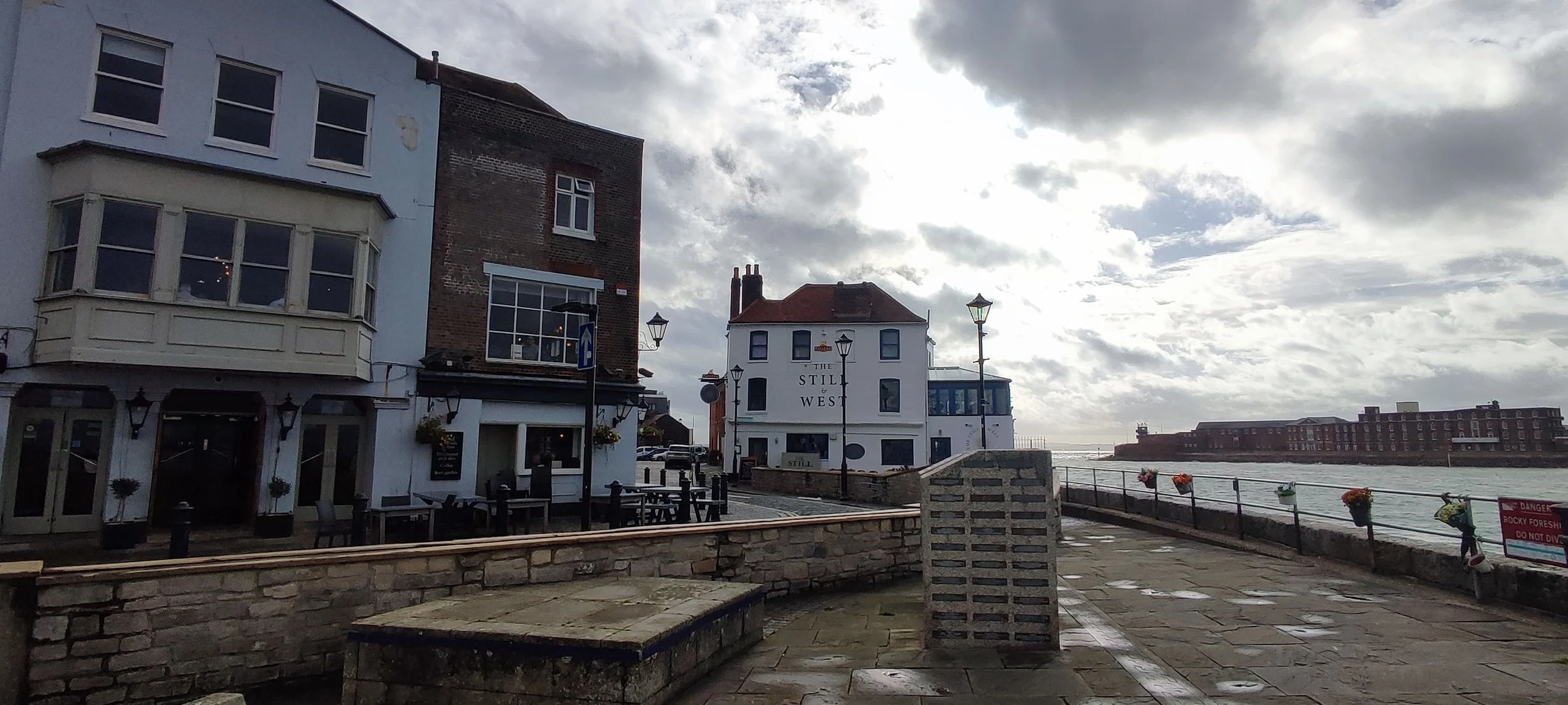 View of a seaside town with buildings along the waterfront, including a white building labeled 'The Still & West,' and a distant large brick building over the water. Cloudy sky overhead.