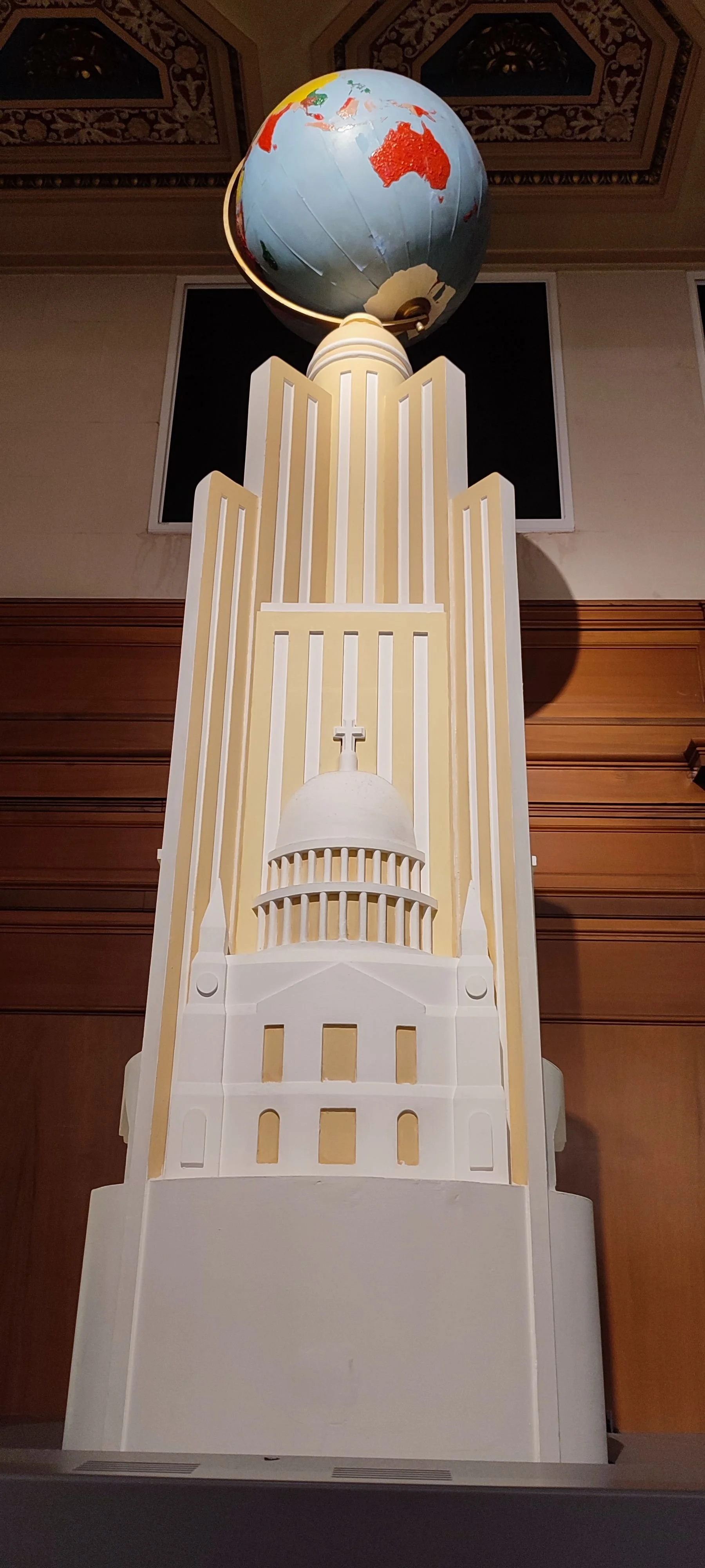 A tall white and yellow architectural model resembling a church with a dome and cross at the top, topped with a globe showing parts of Australia and Asia.