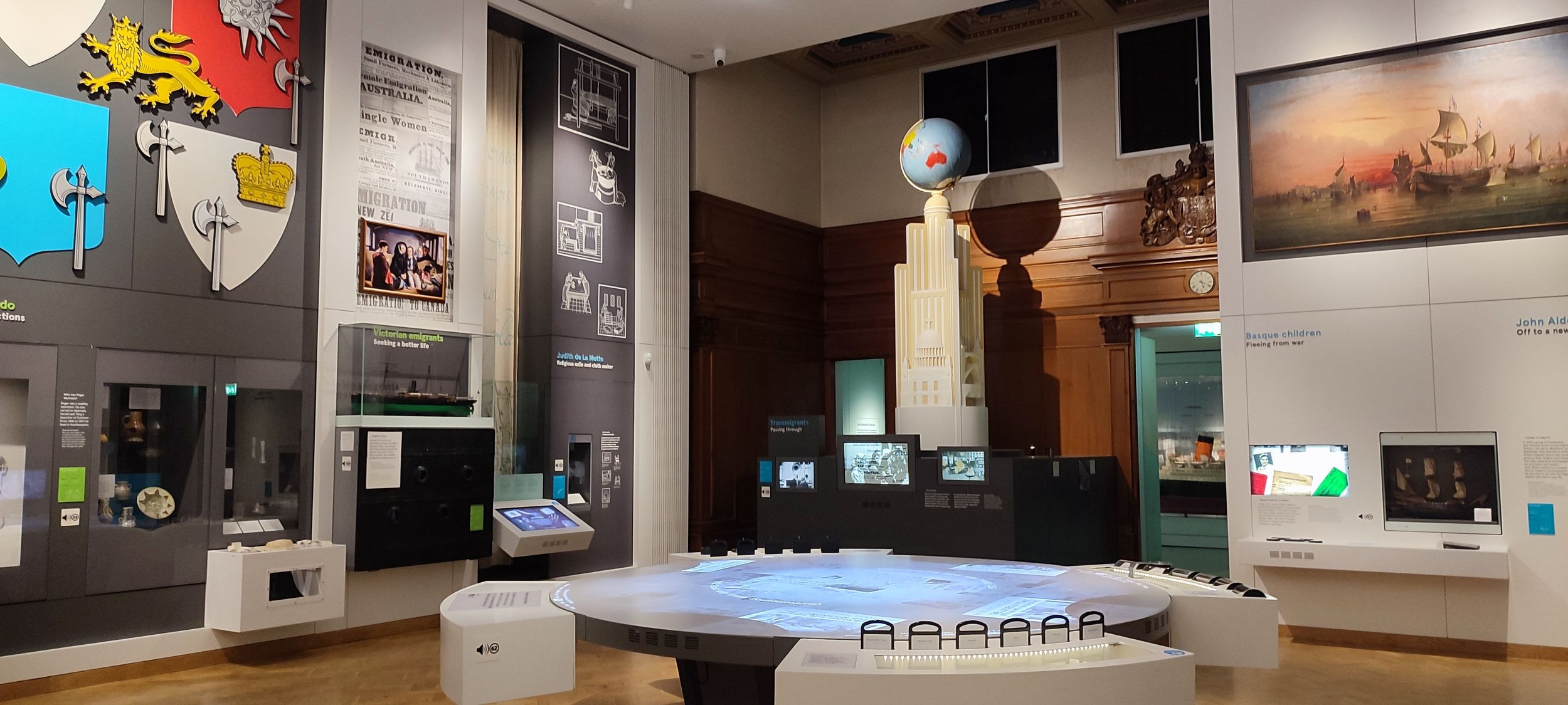 Museum exhibit displaying shields, a ship model, and various informational panels, with a large model of a tower and globe structure in the center, and a painting of ships at sea on the wall.